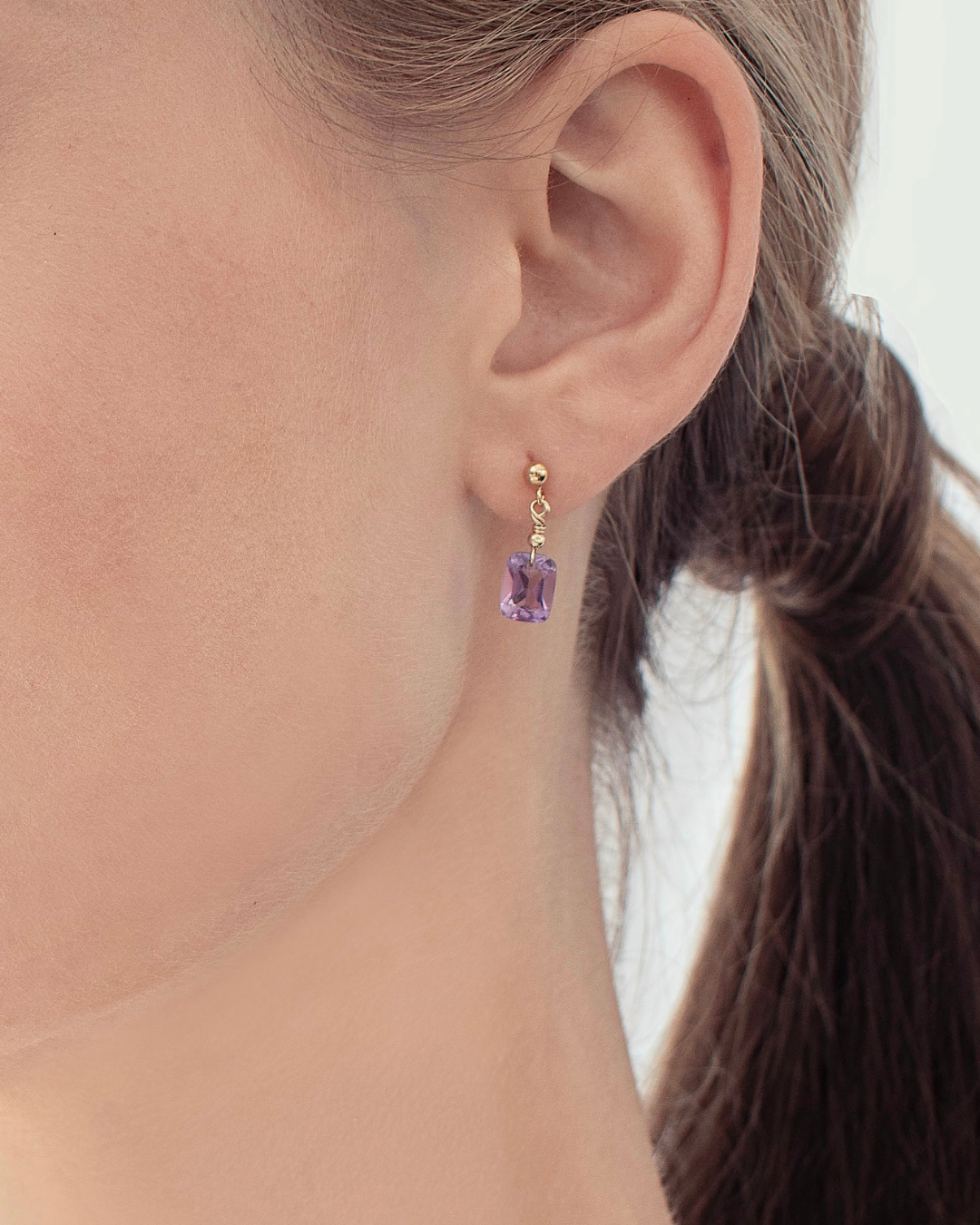 Close-up of an ear wearing a purple amethyst gemstone earring with gold setting.