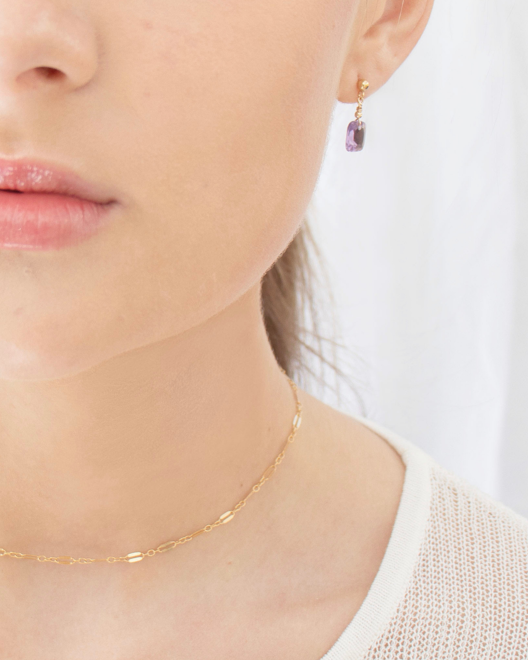 Close-up of a person wearing purple amethyst gold earrings and a gold choker necklace with a blurred background