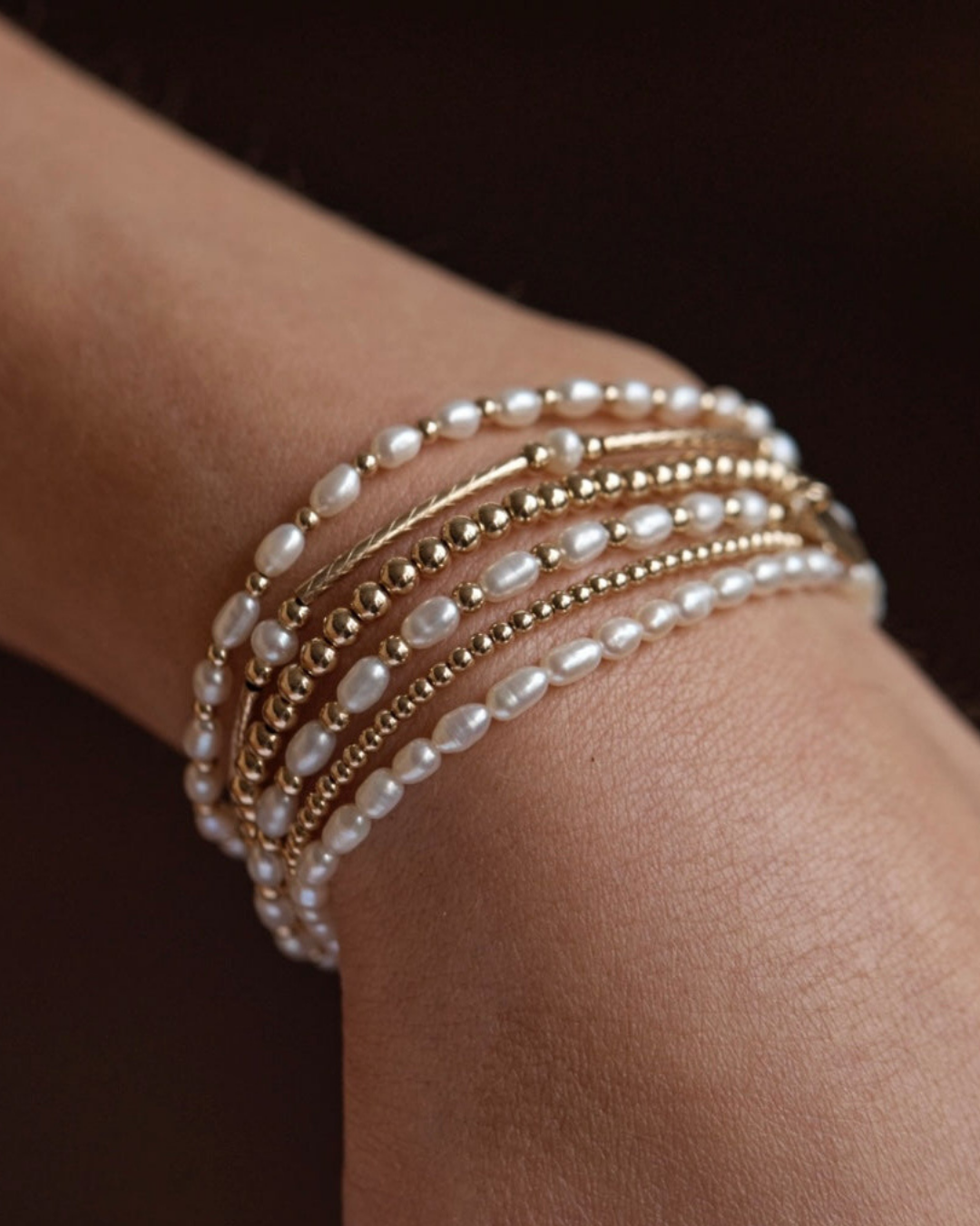 Close up of multiple pearl and gold bracelets on a wrist against a dark background