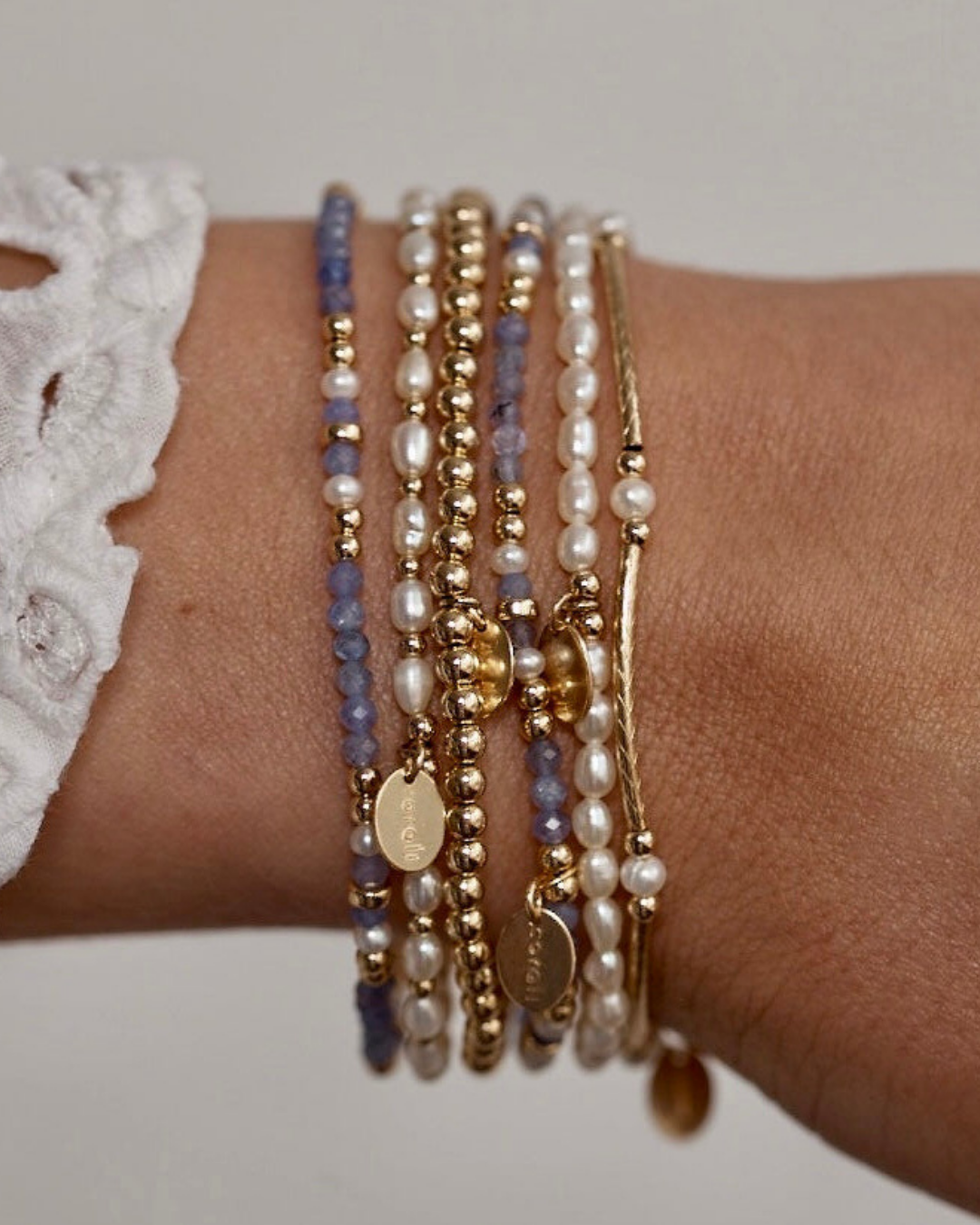 Close-up of a wrist wearing multiple beaded gold pearl and gemstone bracelets on a neutral background