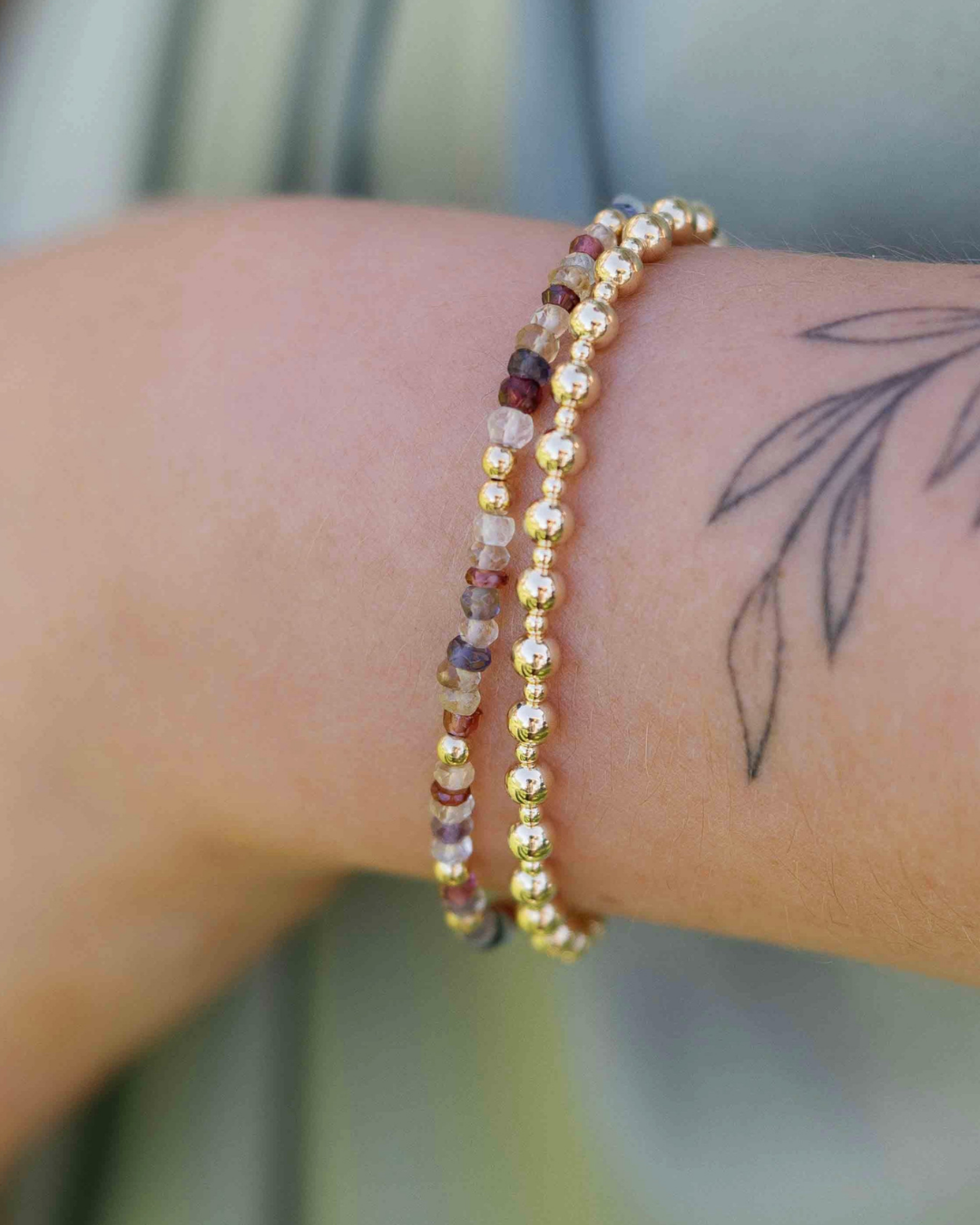 Close-up of a wrist wearing two gold beaded bracelets with a tattoo on the skin.