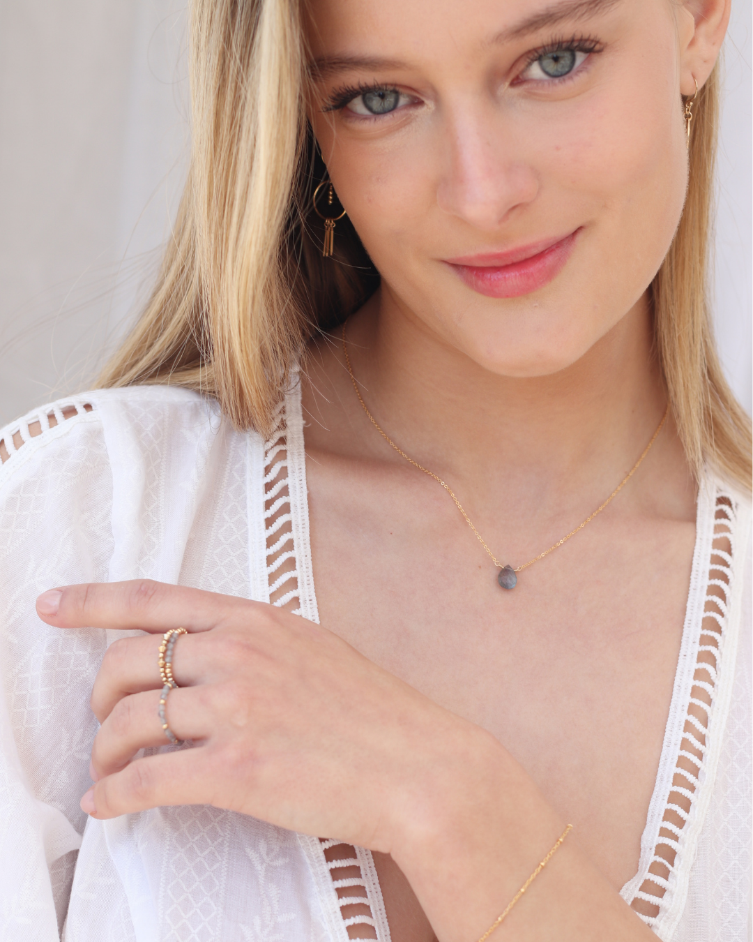 Woman wearing a delicate necklace with a  teardrop-shaped labradorite pendant on a gold chain against a white background and bracelet, gold rings, and earrings.