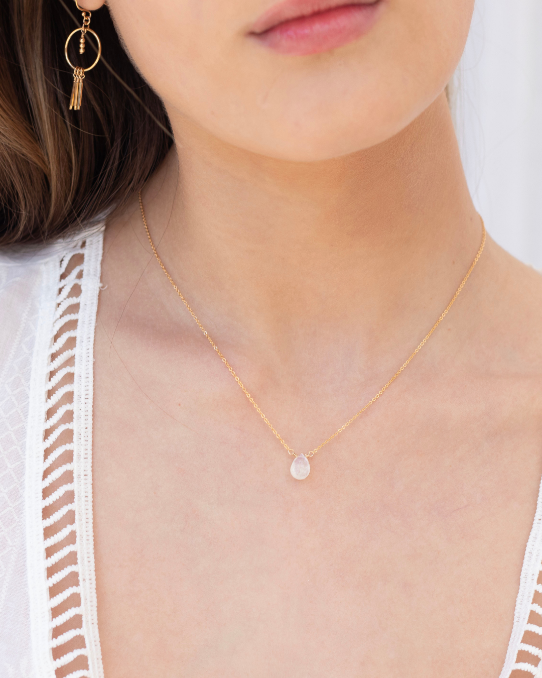 Close-up of a person wearing a gold necklace with a tear drop moonstone small pendant and gold earrings.