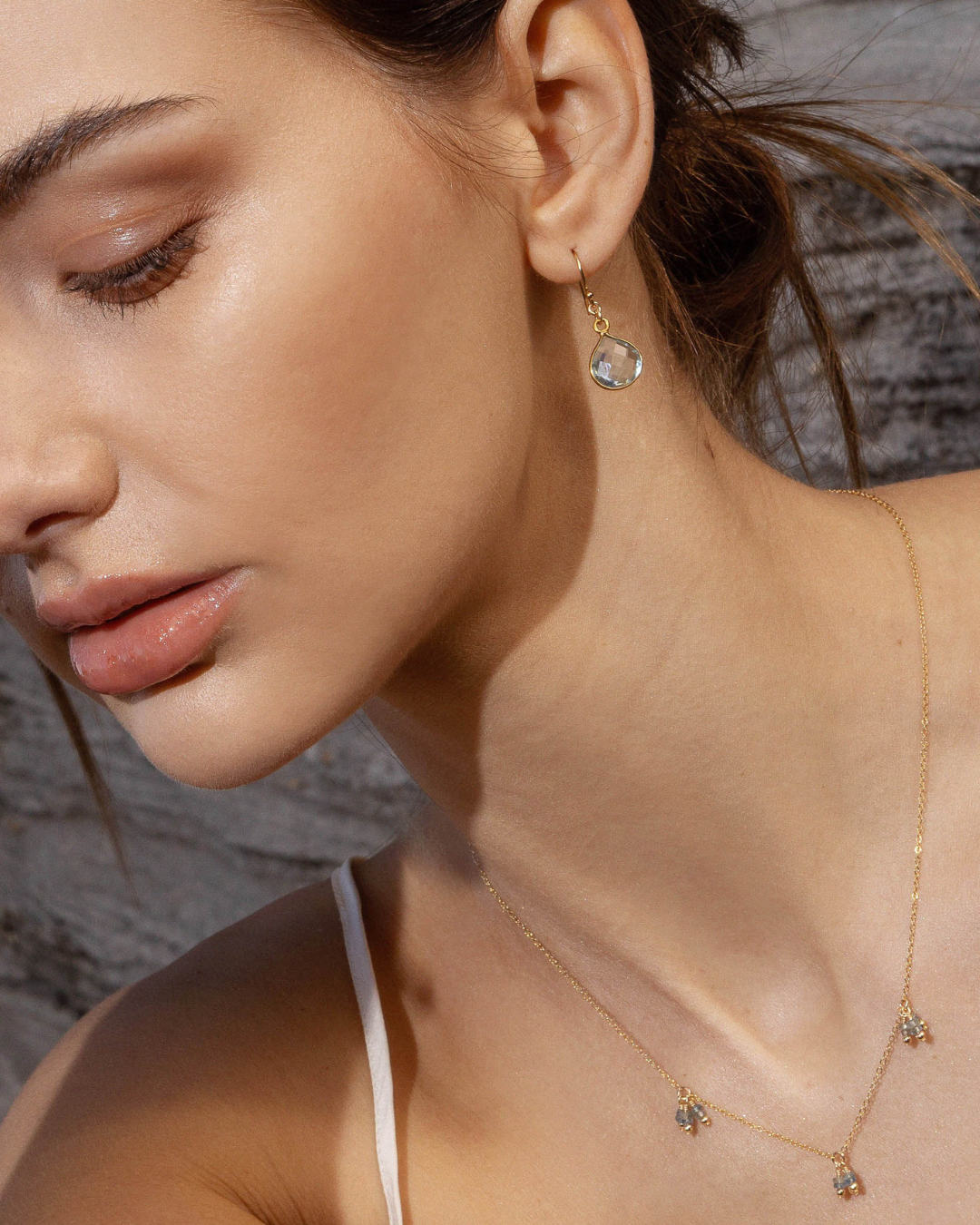 Close-up of a woman wearing gold earrings with blue topaz gemstone and necklace with aquamarine gemstones against a stone wall background