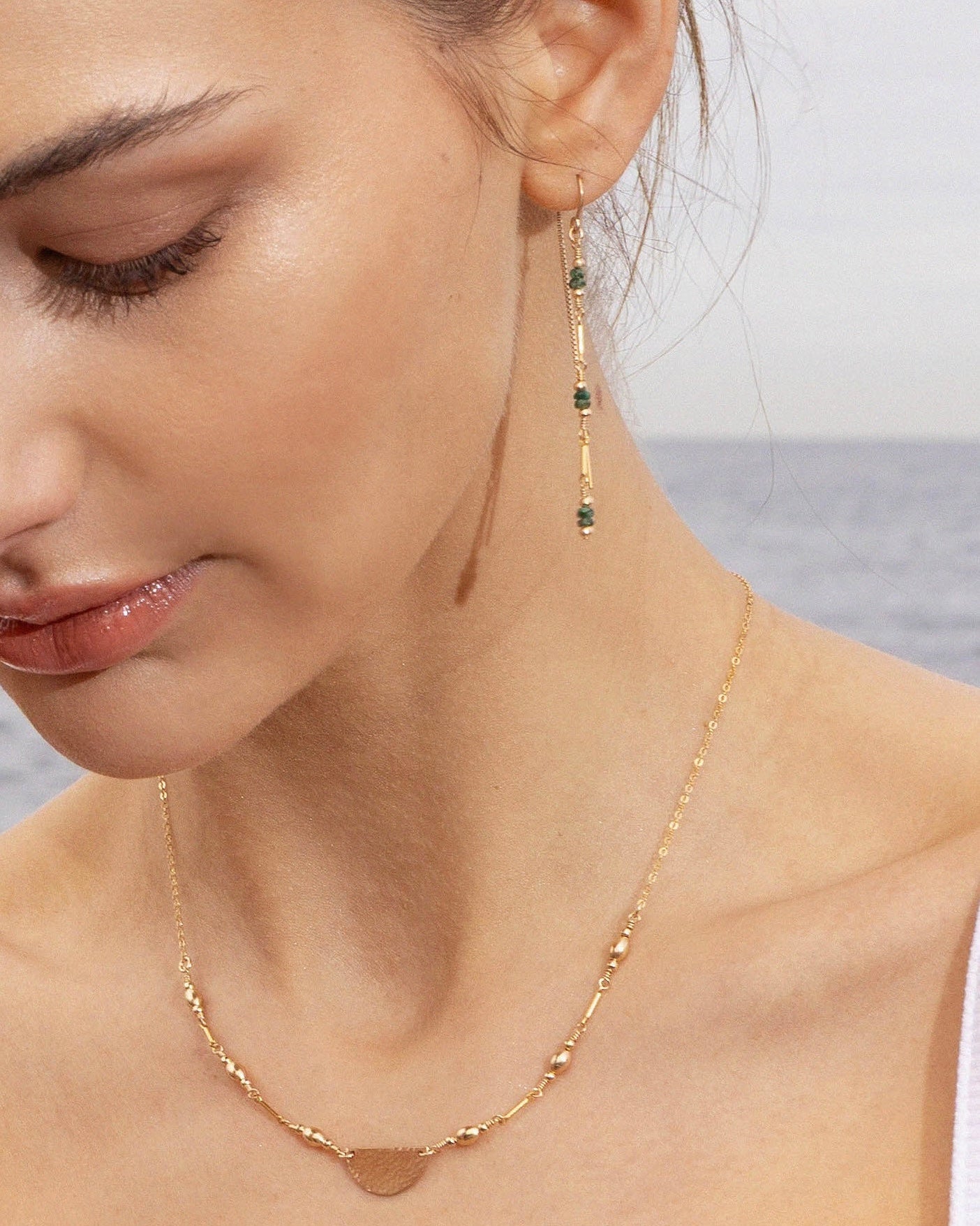 Close-up of a woman wearing a gold thread earring with emerald gemstones and a gold necklace with a blurred ocean background