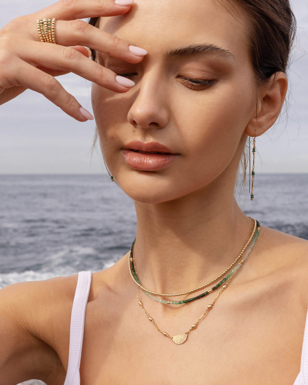Woman wearing layered gold and emerald gemstone necklaces and green emerald gemstone gold thread earrings with a blurred ocean background