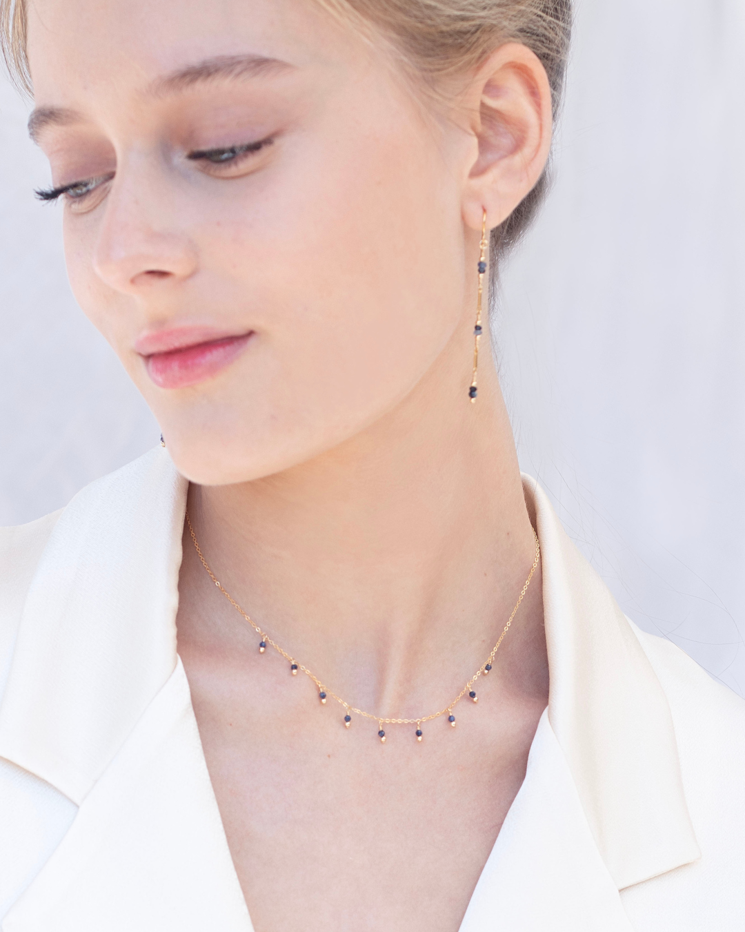 Close-up of a person wearing gold and sapphire thread earrings with a delicate necklace with small sapphire gemstones on a plain background