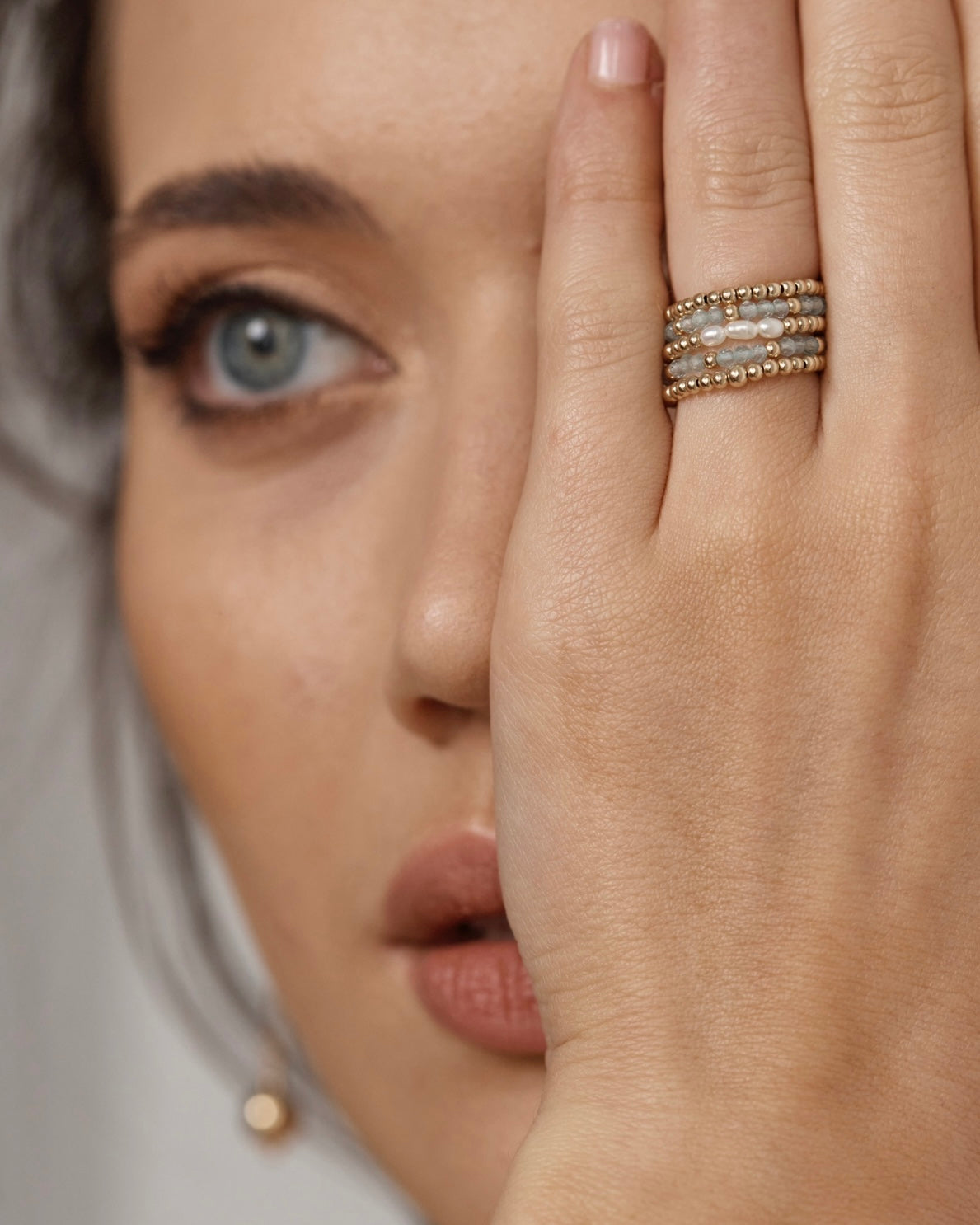 Close-up of a woman's face with one hand covering her eye, wearing gold beaded apatite gemstone rings, gold bead rings and a gold ring with pearls.