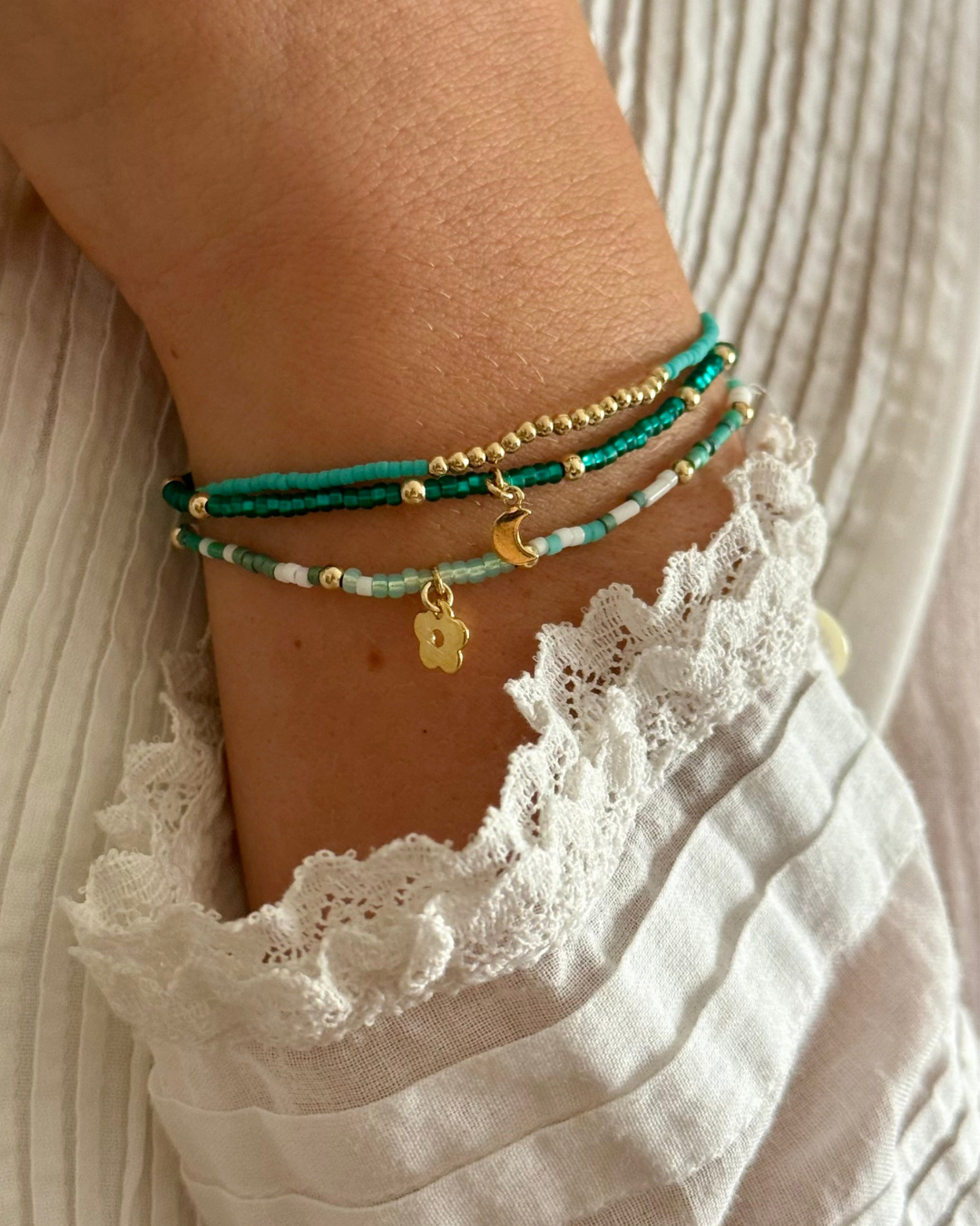 Close-up of a wrist wearing multiple glass seed turquoise, teal, white and gold beaded bracelets with a white lace garment.
