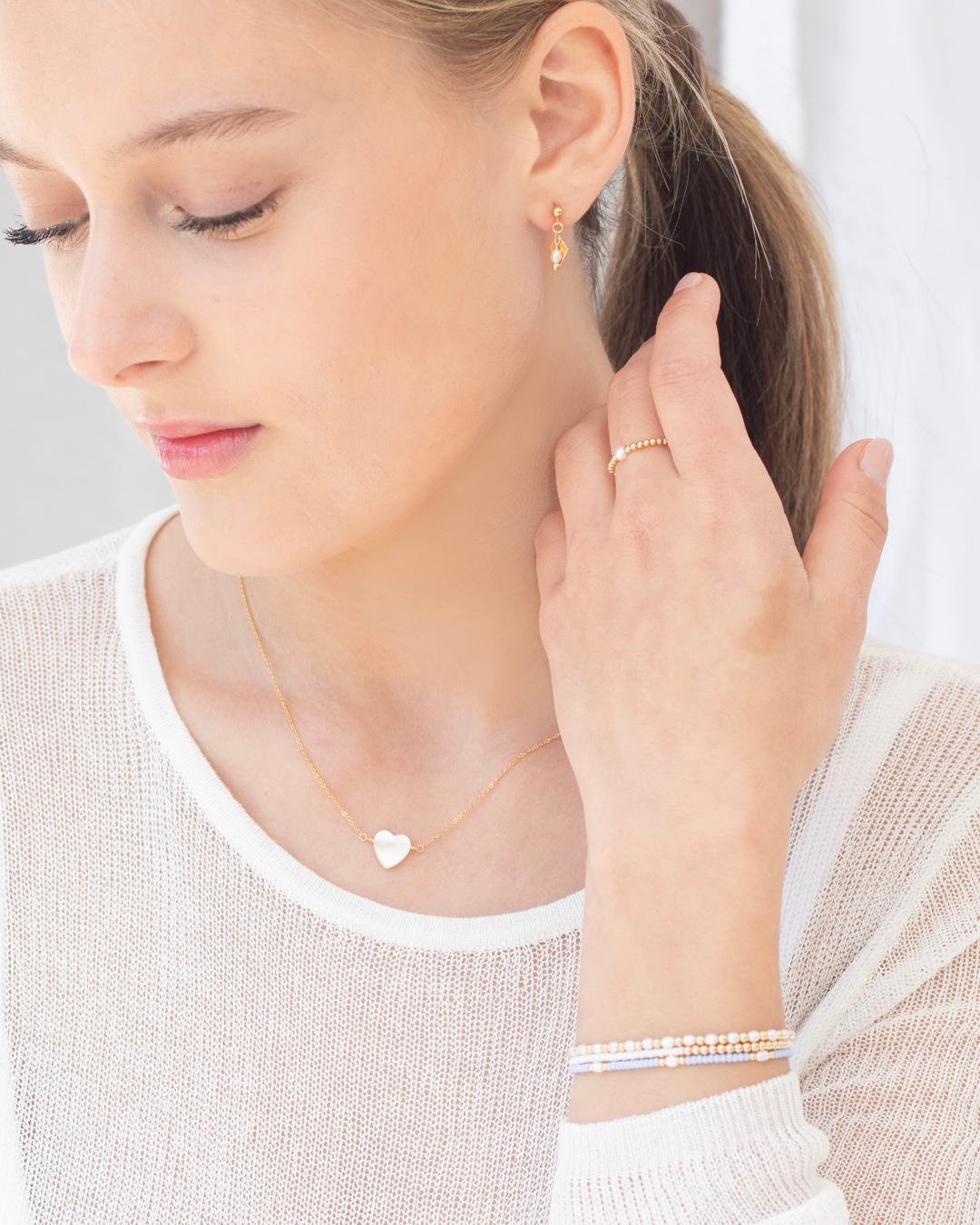 Woman wearing a heart-shaped mother of pearl pendant necklace, pearl, earrings, ring and bracelets on a plain background