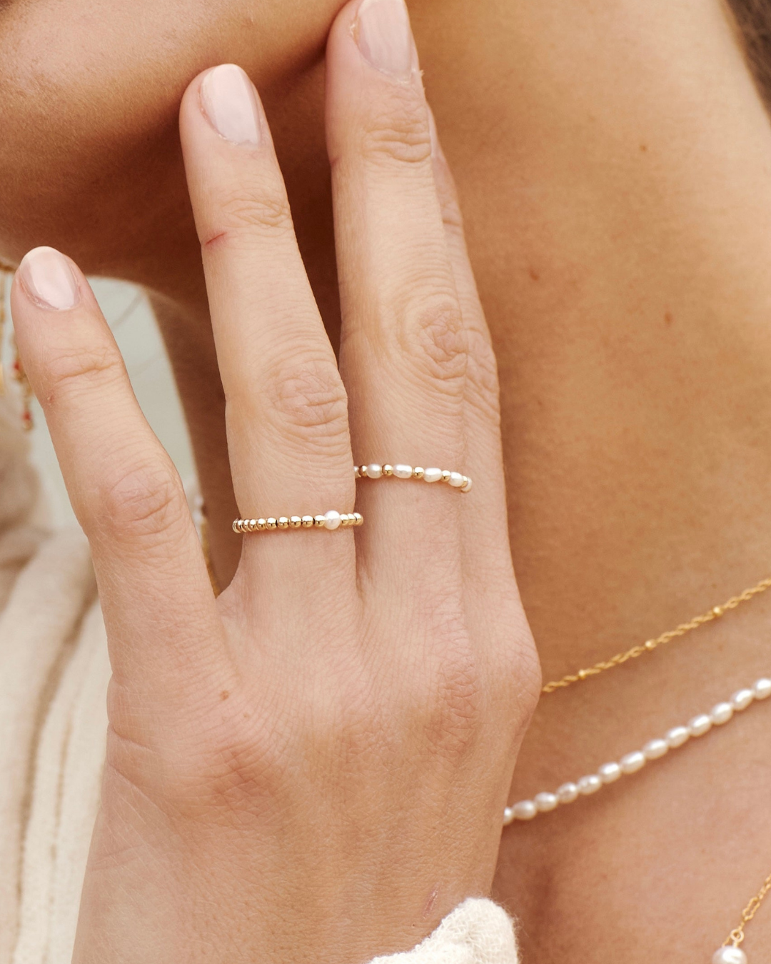 Close-up of a hand wearing two gold and pearl rings with a pearl necklace.