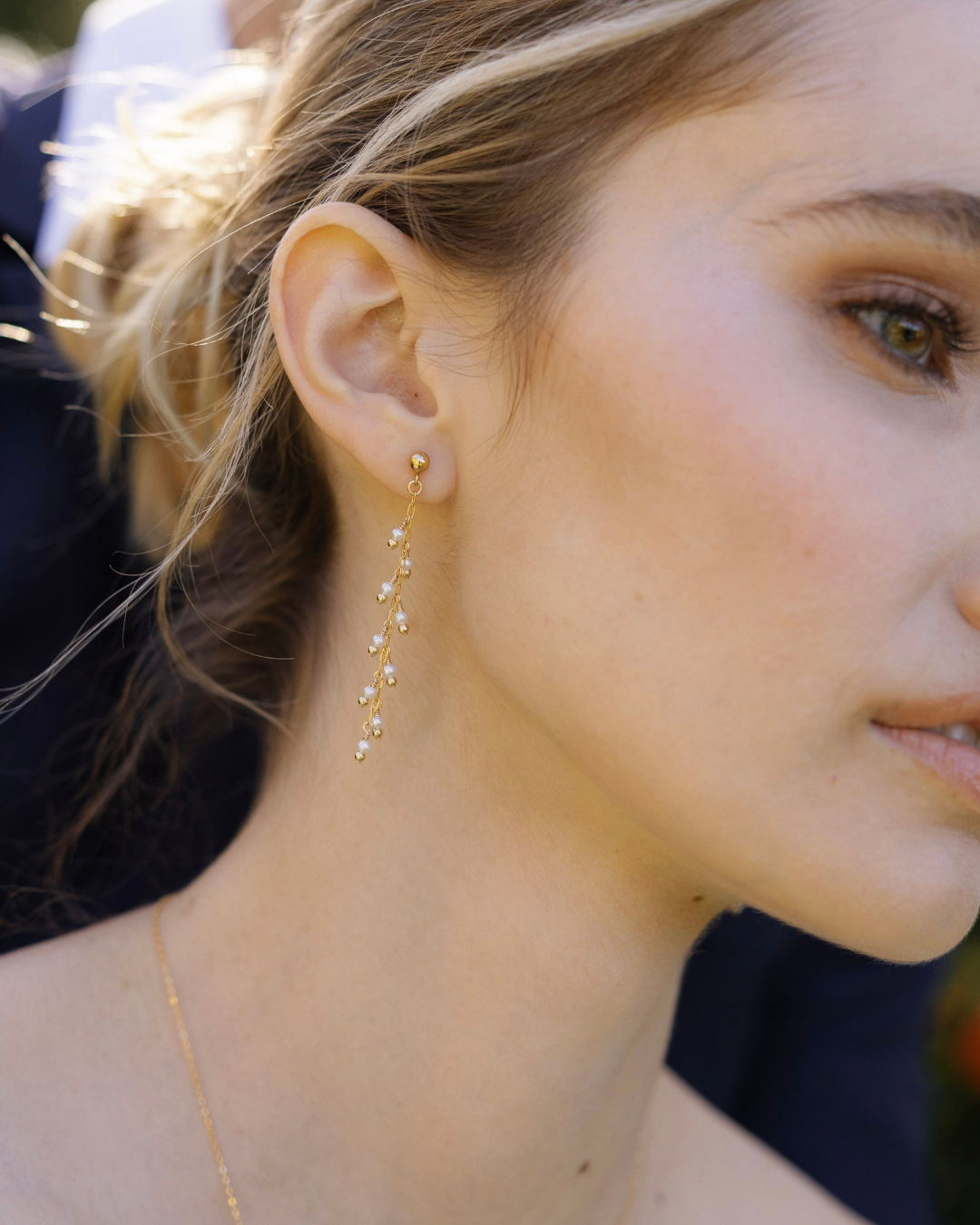 Close-up of a bride wearing gold and pearl earrings and a necklace outdoors.