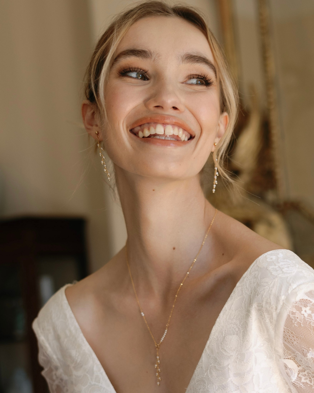 Bride wearing a gold and pearl necklace and earrings, smiling in a softly blurred indoor setting