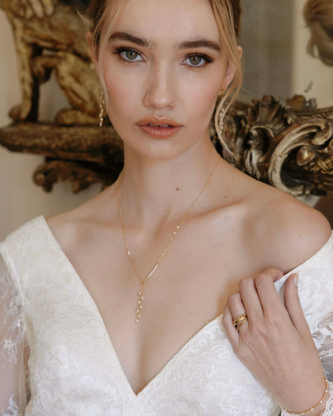 Bride wearing a gold and pearl necklace, earrings and rings in front of an ornate gold mirror.
