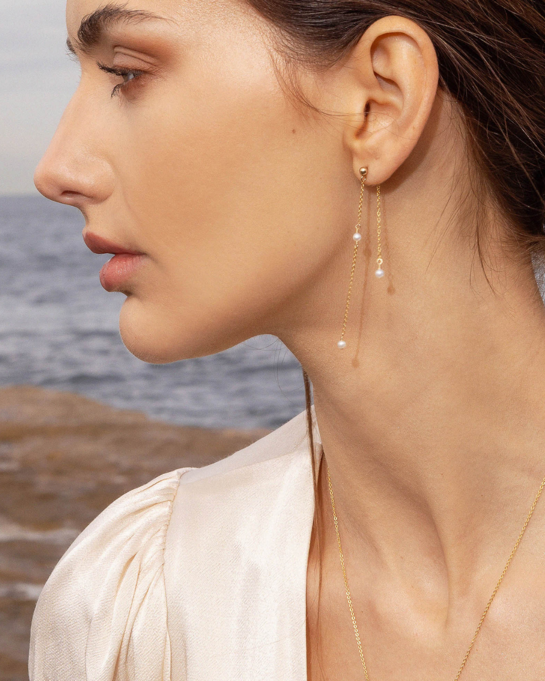 Close-up of a woman wearing gold pearl earrings with a blurred ocean background