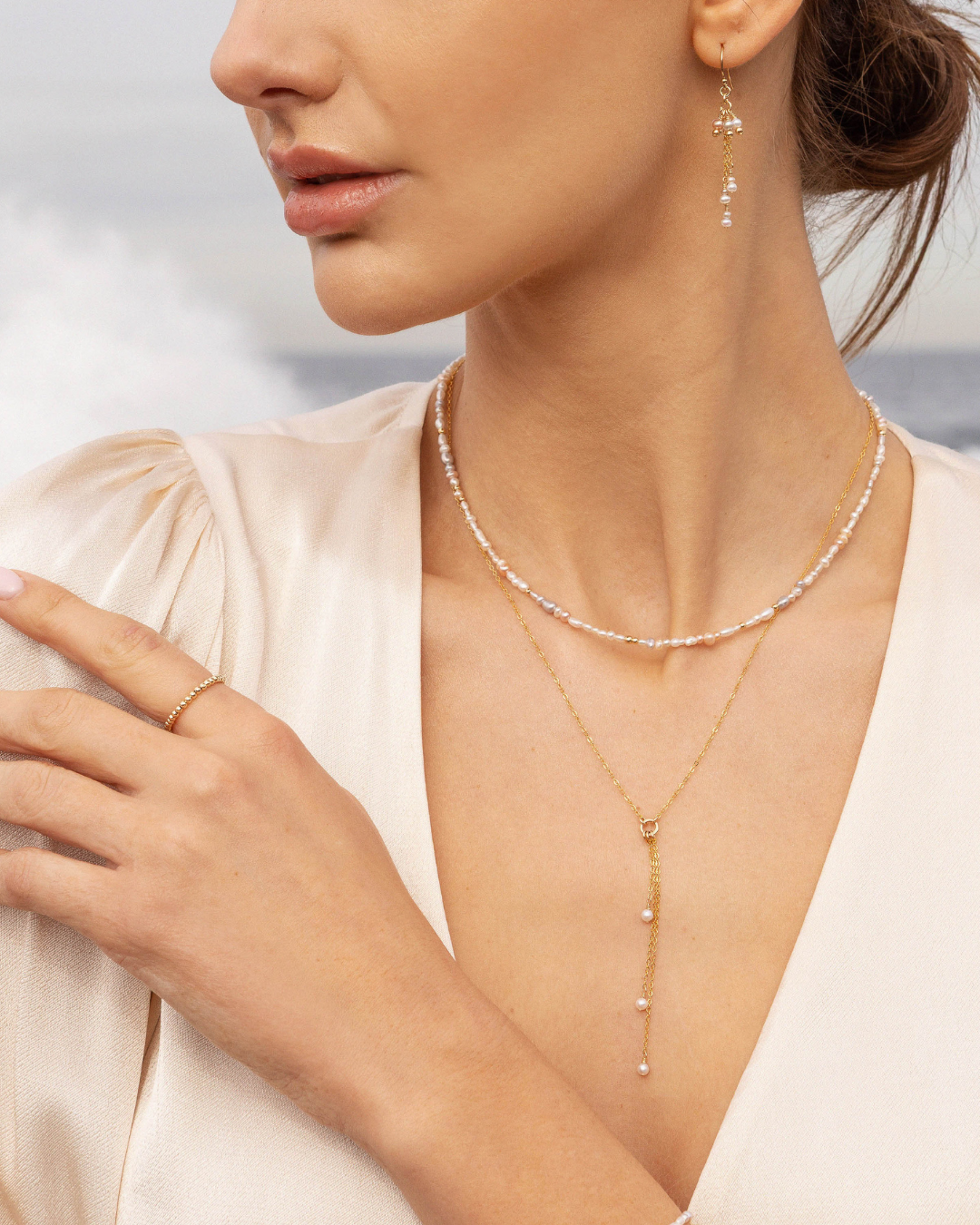 Woman wearing a delicate gold pearl lariat necklace, ring and earrings with a blurred background