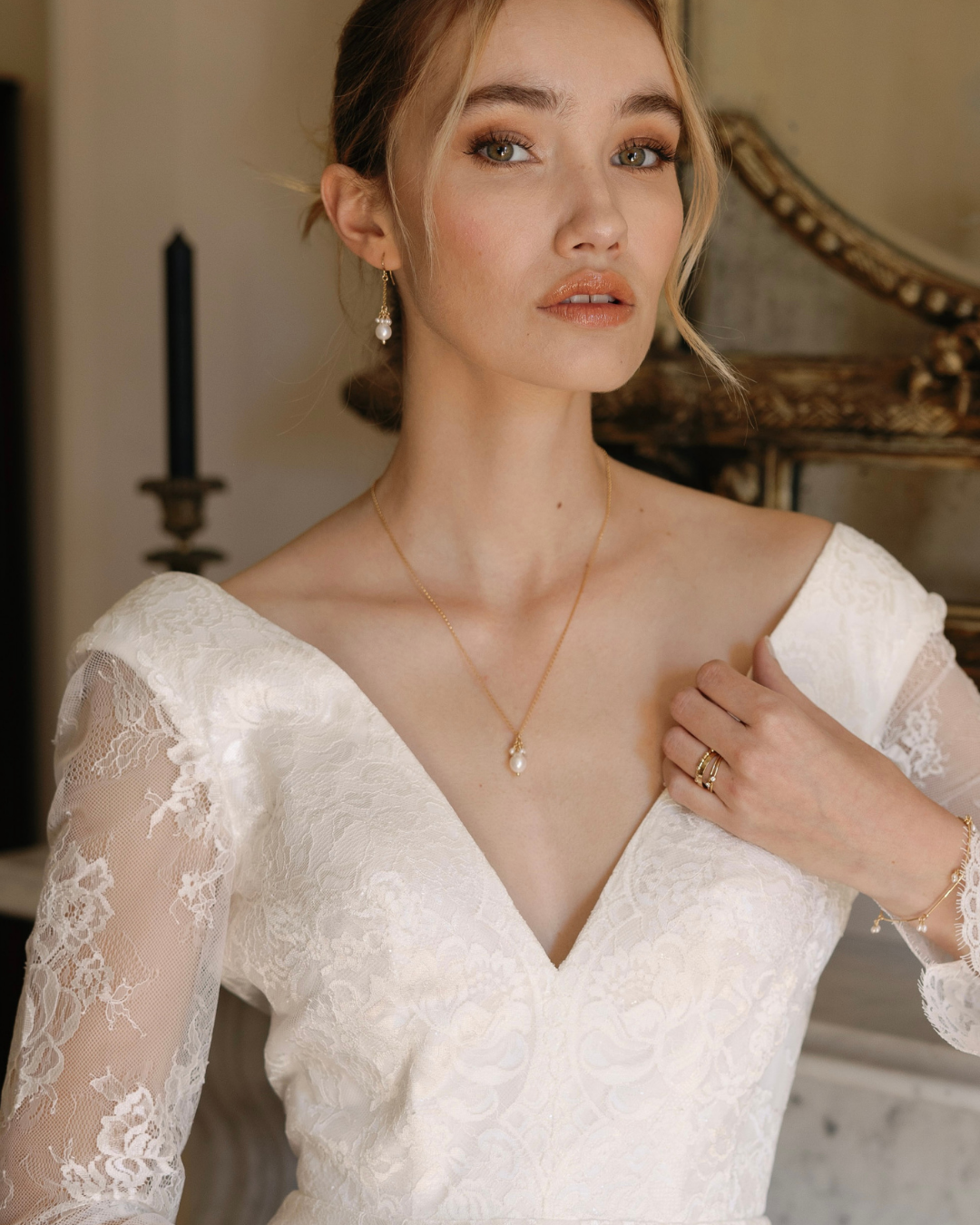 Woman wearing a white lace dress with a gold necklace and ring in an elegant indoor setting.