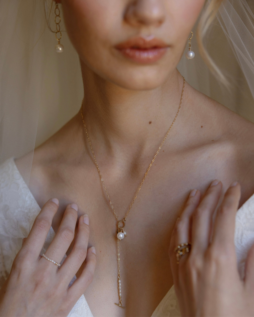 Close-up of a bride wearing a gold lariat necklace with a pearl pendant, earrings, and rings.