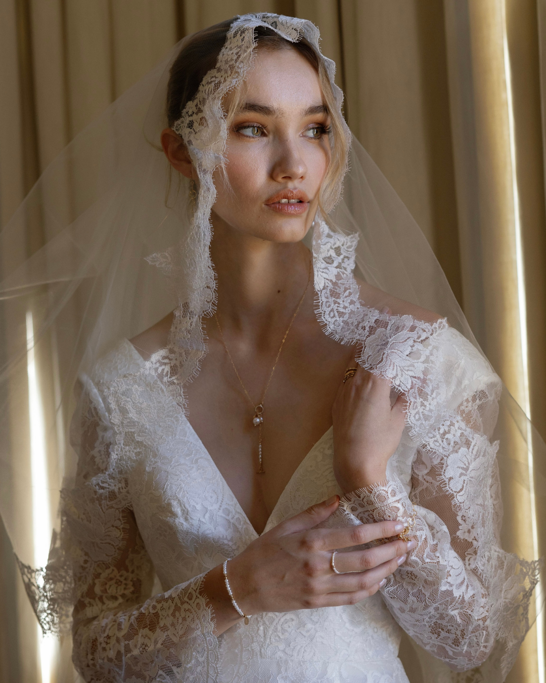 Bride wearing a lace veil and wedding dress, with gold and pearl jewellery against a neutral background