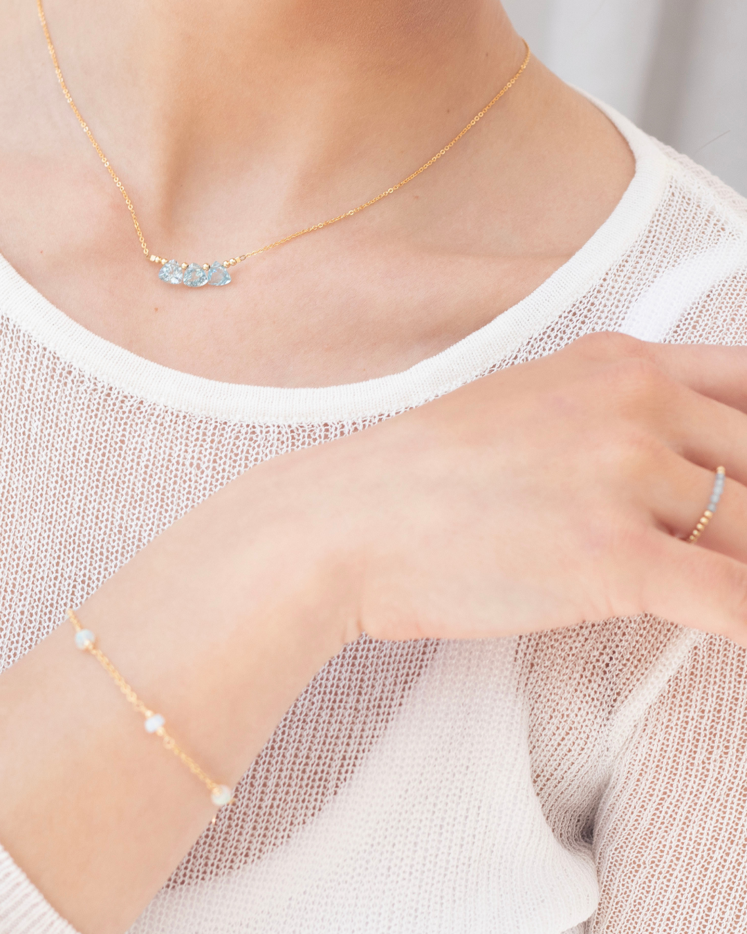 Gold necklace with blue topaz briolette gemstones, opal bracelet, and a larimar gemstone beaded ring on a woman wearing a white top.