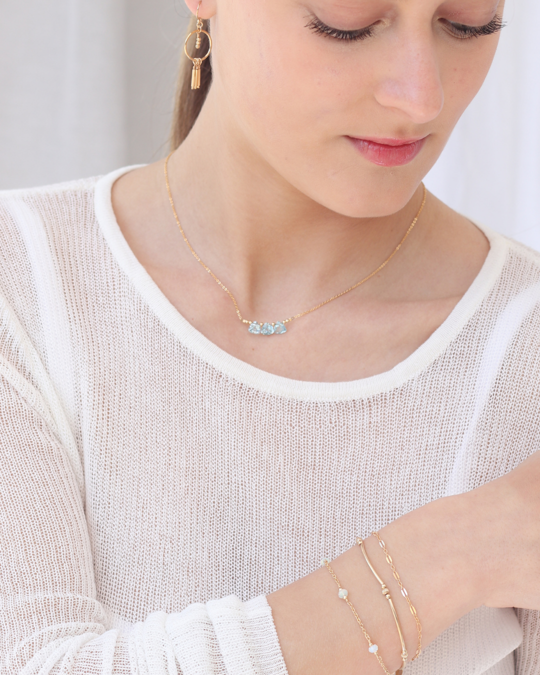 Woman wearing a white top featuring a gold necklace with blue topaz briolette gemstones, layered bracelets, and a pair gold of earrings.