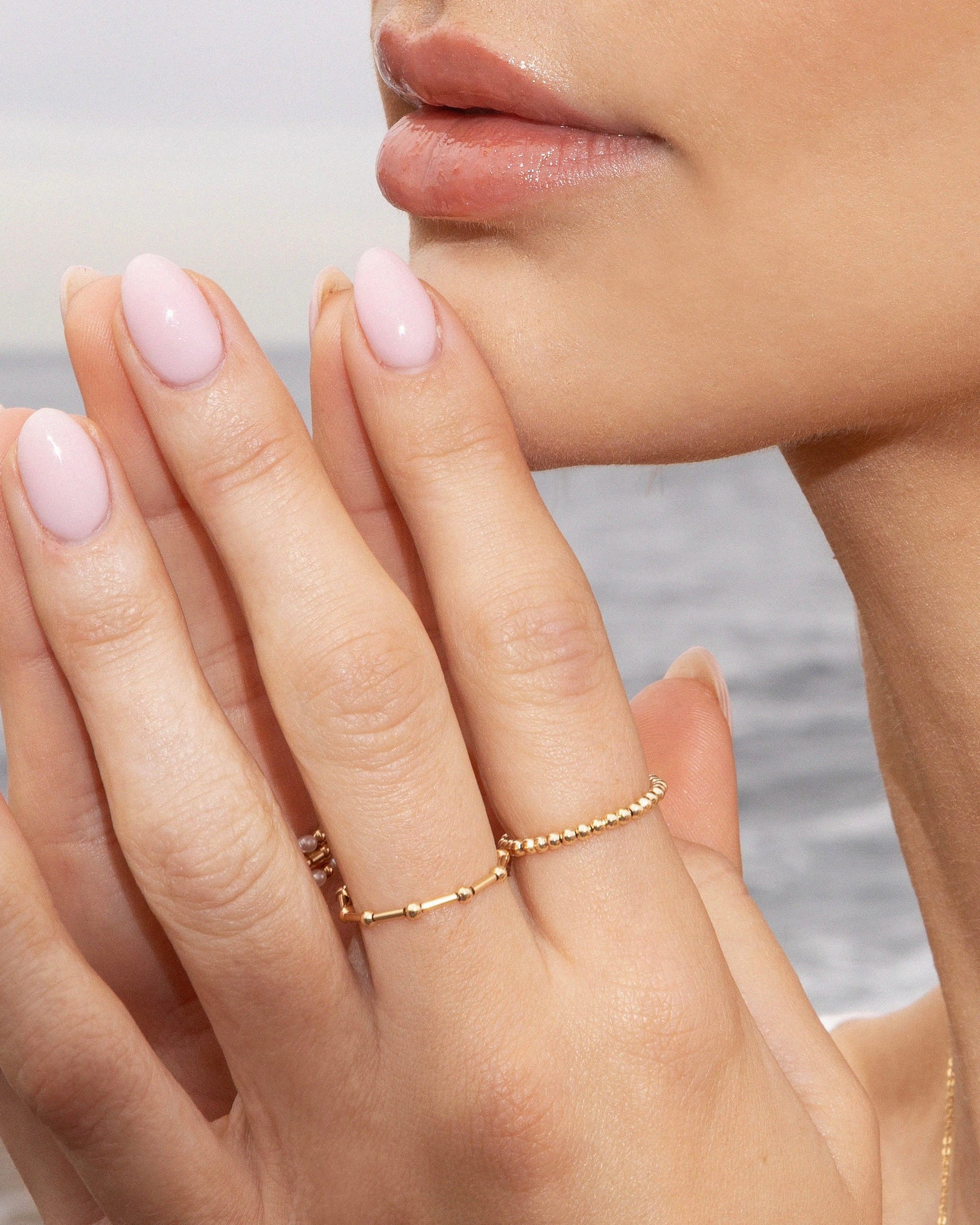 Close-up of a woman's hand with pink nail polish and gold rings, against a blurred natural background.
