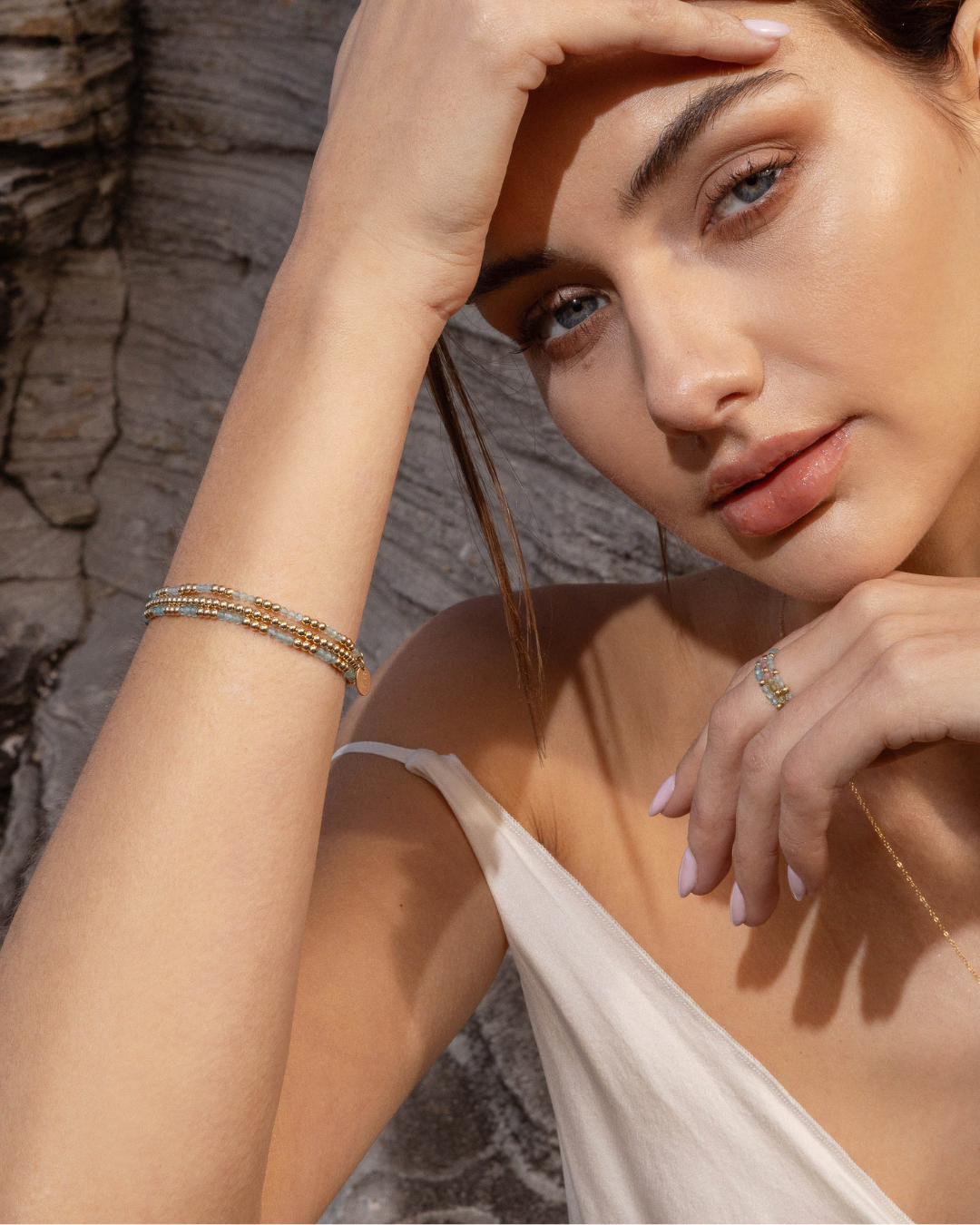 Woman wearing gold bead and apatite gemstone bracelets and rings with a stone wall background