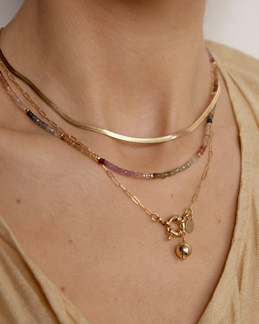 Close-up of a person wearing a multi coloured sapphire gemstone beaded necklace layered with multiple gold necklaces with a neutral background