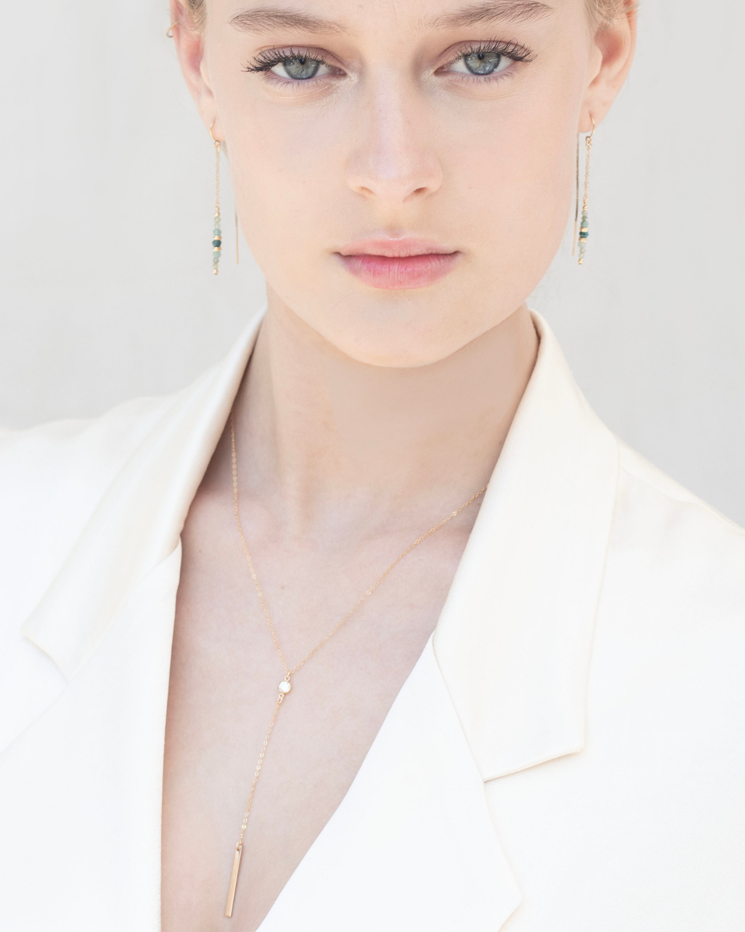 Close-up of a person wearing a delicate gold lariat necklace and earrings on a plain background