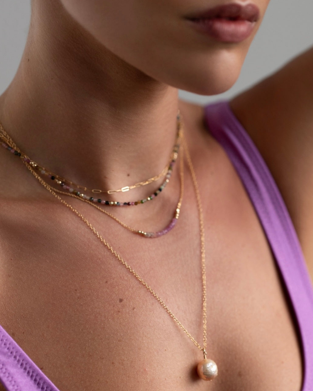 Close-up of a person wearing multiple gold necklaces with a focus on one featuring a tourmaline gemstone necklace.