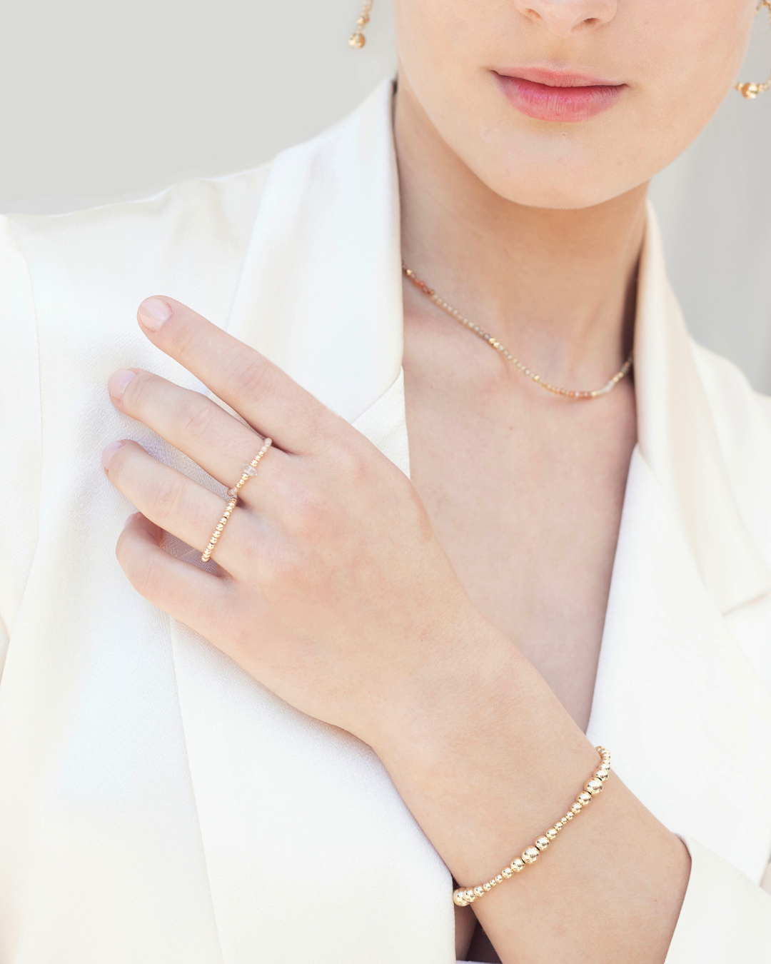 Close-up of a woman wearing gold jewellery including rings, a bracelet, and zircon necklace on a plain background.