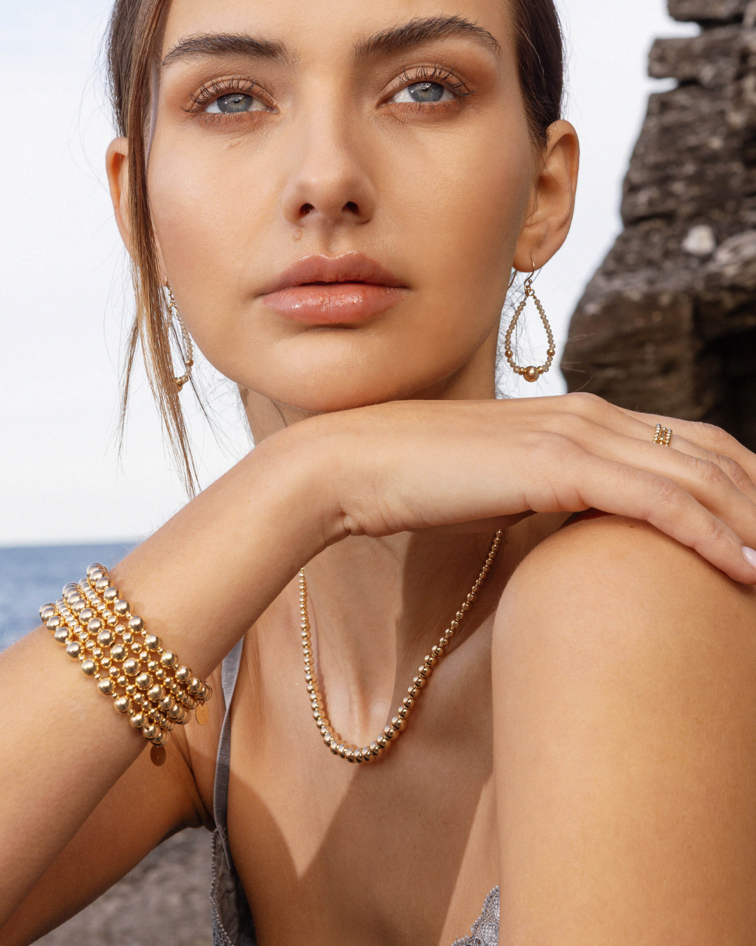 Woman wearing gold jewellery including a necklace, bracelets, rings, and zircon gemstone earrings with an ocean background.