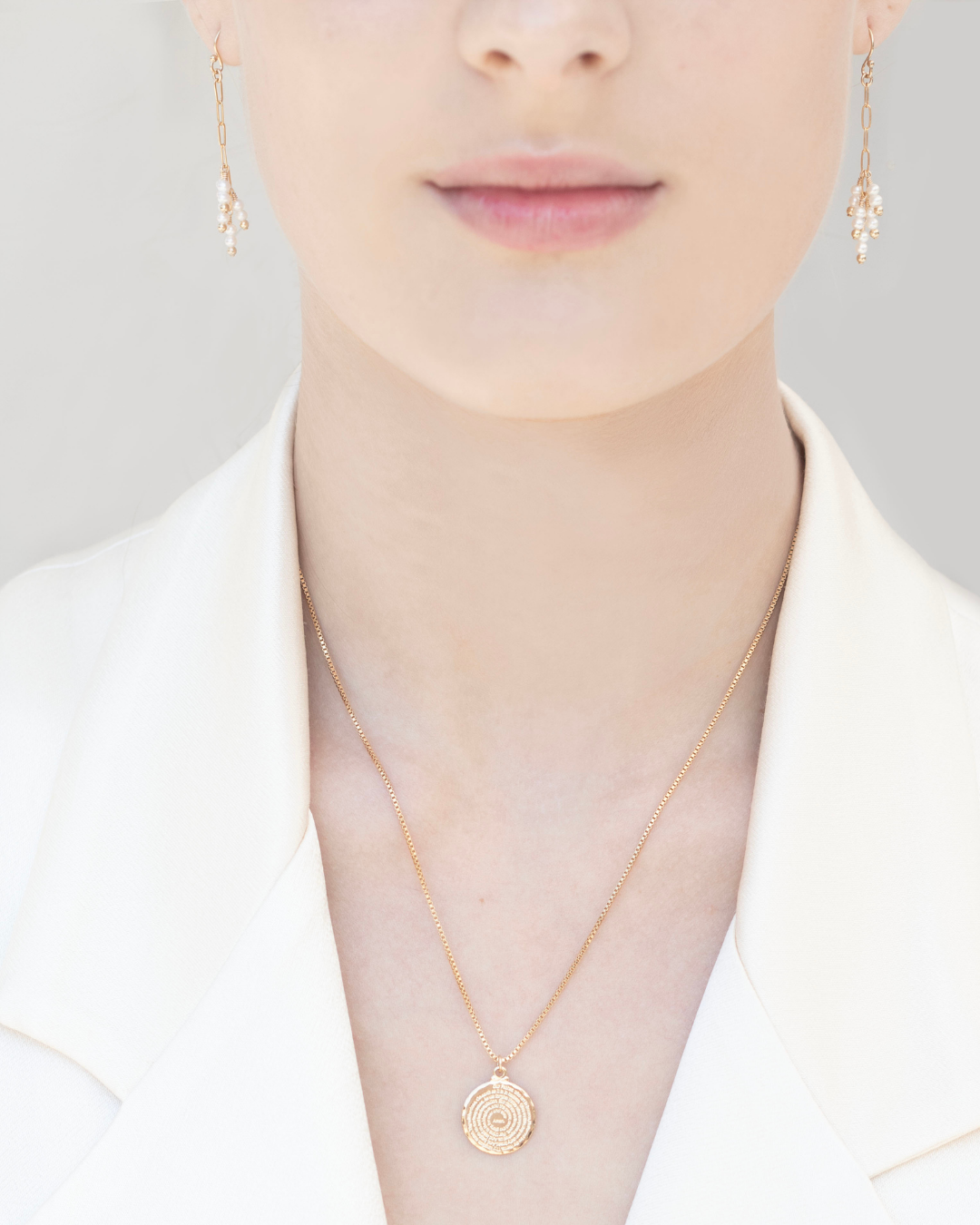Close up of a woman wearing gold pearl earrings and a gold pendant necklace against a neutral background