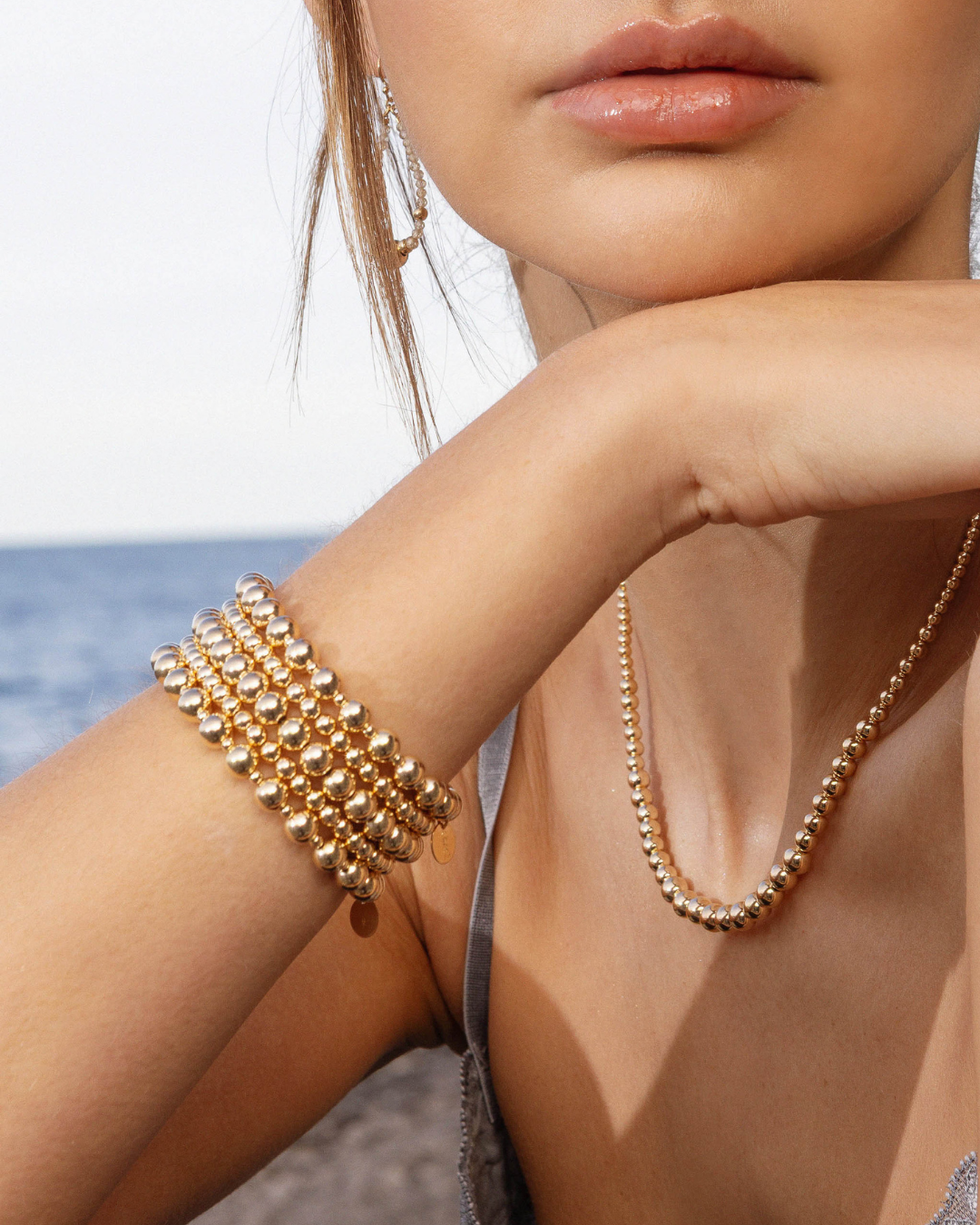 Close-up of a person wearing gold beaded bracelets and necklace with an ocean background