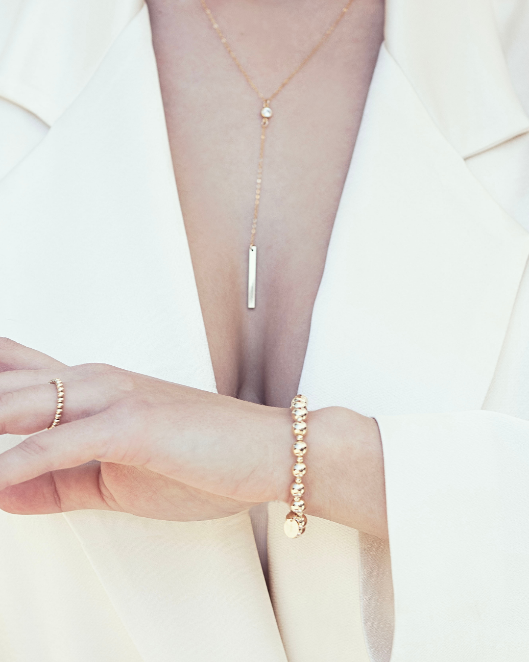 Model wearing a gold bead bracelet, ring and fine gold lariat cubic zirconia gemstone necklace, wearing a  cream satin outfit.