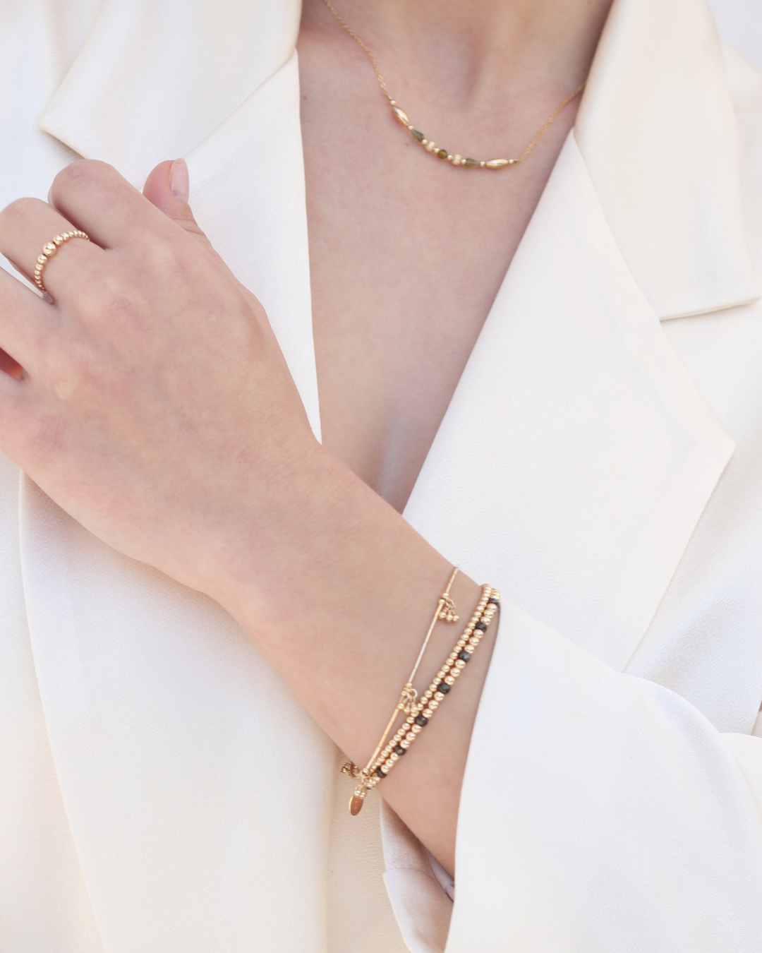 Close-up of a person wearing gold jewellery featuring a green zircon bracelet stacked with gold bead bracelets, a ring, and a green tourmaline gemstone necklace on a white background.