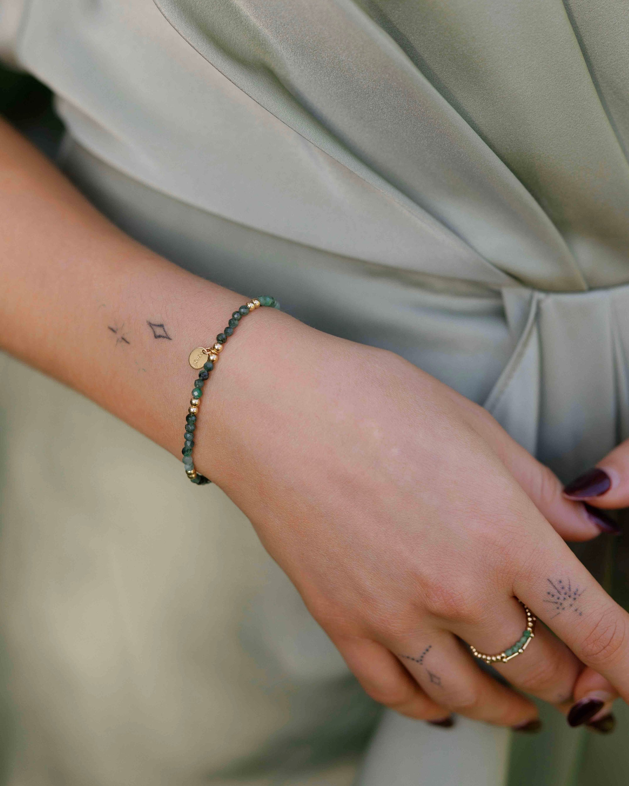 Close-up of a woman's wrist wearing an emerald bracelet and rings, wearing a green silk dress. 