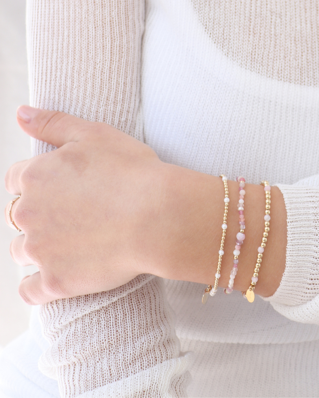Wrist wearing multiple gold beaded pearl and pink gemstone bracelets on a neutral background