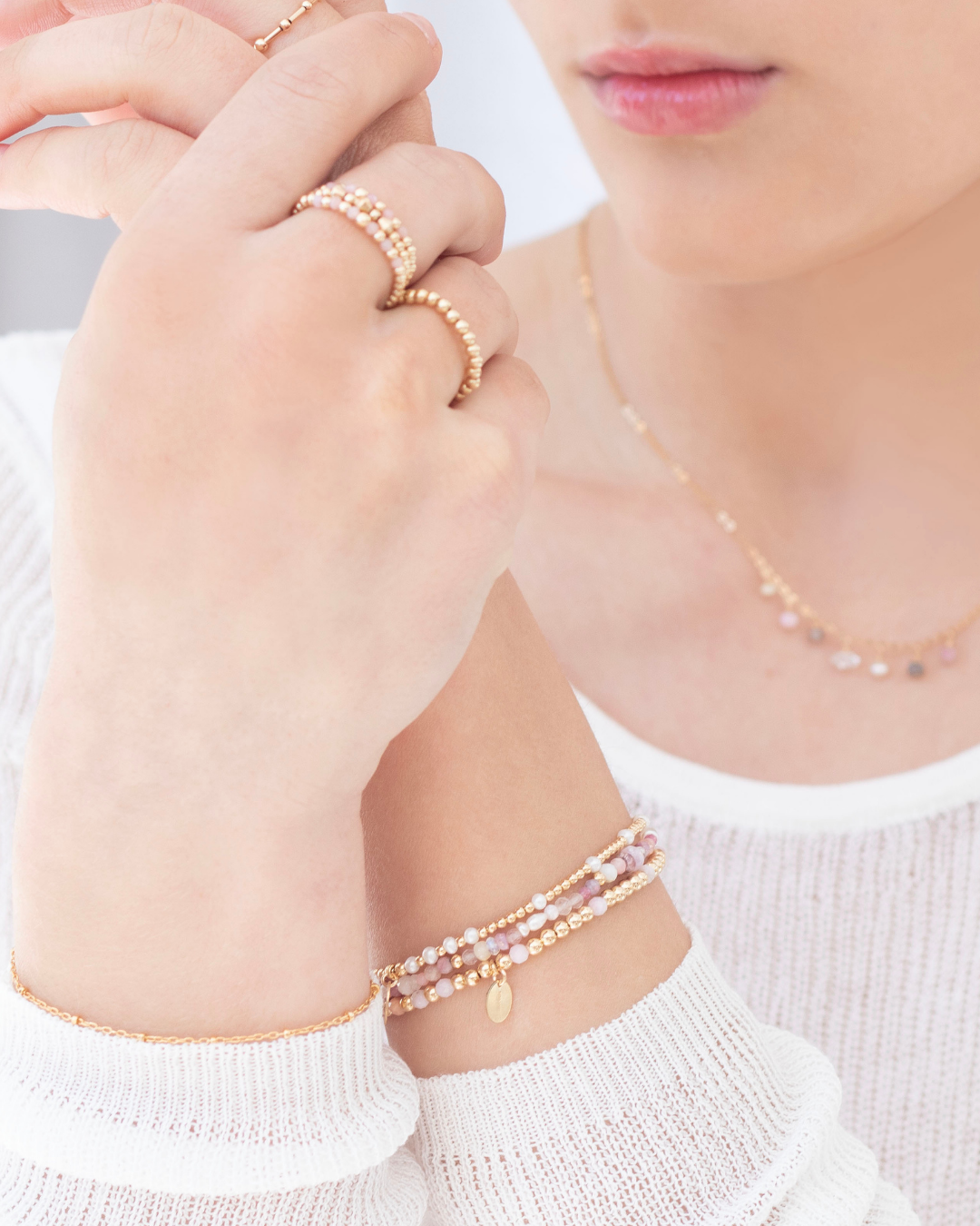 Close-up of a model wearing gold rings, bracelets, and necklace with pearls and pink gemstones wearing a white top