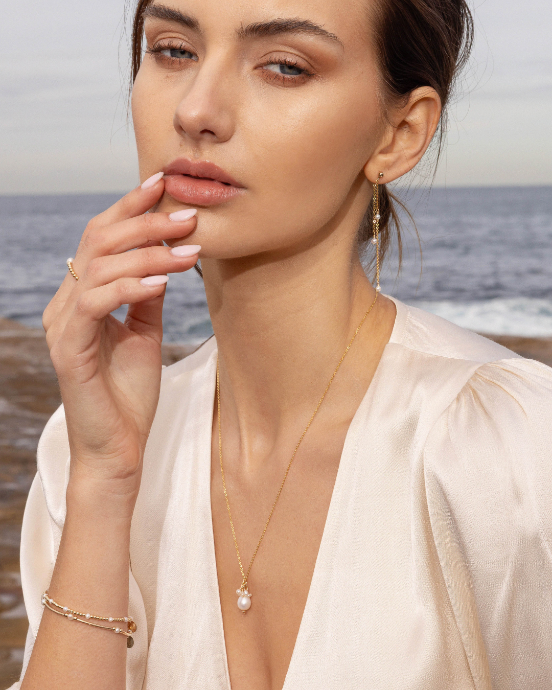Woman wearing gold pearl jewellery with a bracelet, necklace and earrings by the ocean.