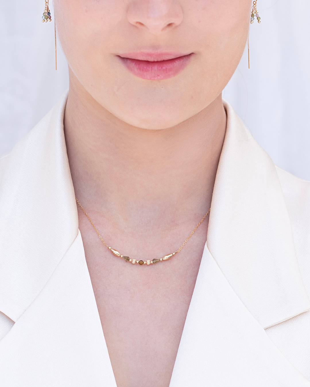 Woman wearing a delicate gold green tourmaline necklace and tourmaline earrings wearing a cream satin blazer with a plain background