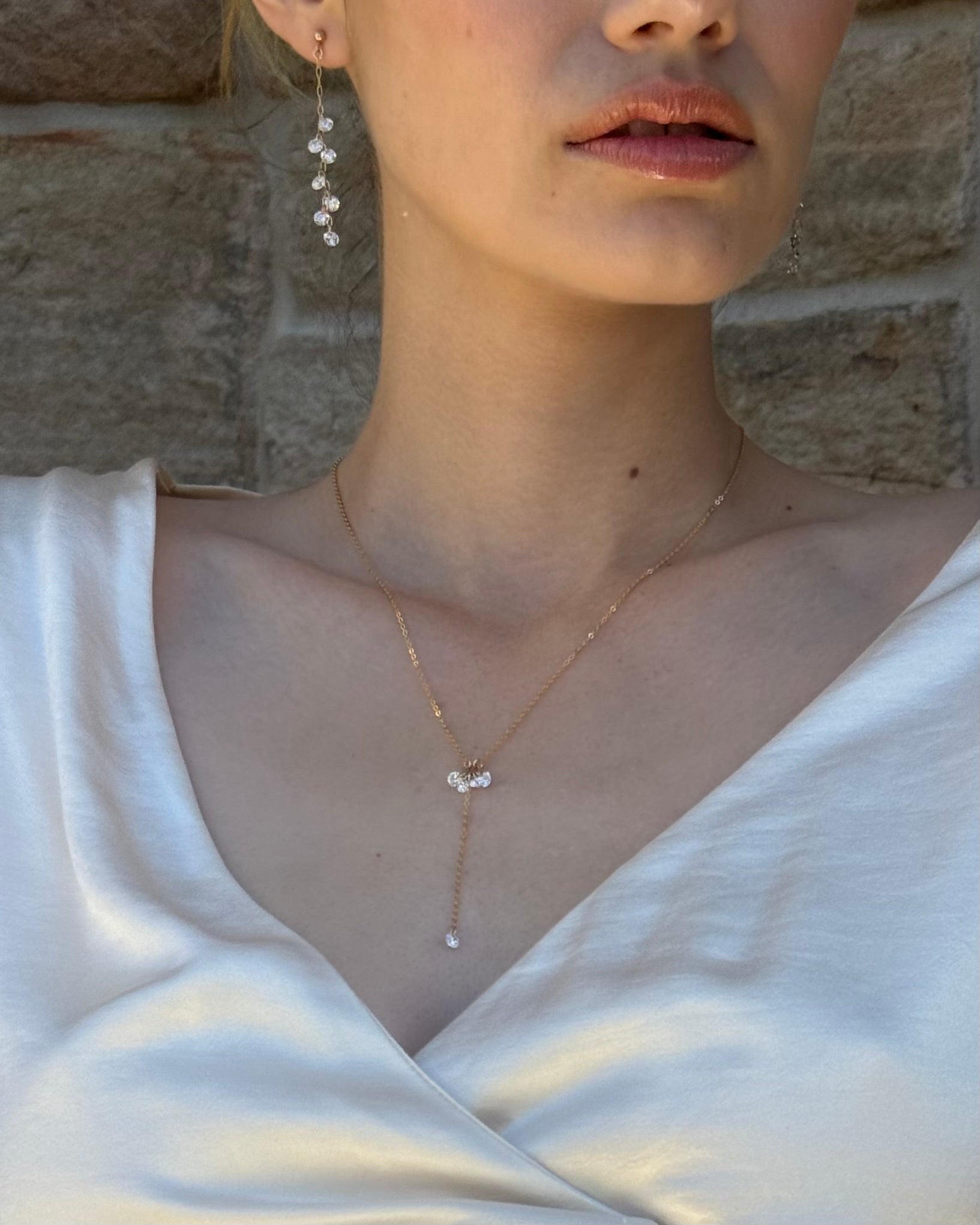 Close-up of a woman wearing a delicate cubic zirconia lariat necklace and earrings against a stone wall.