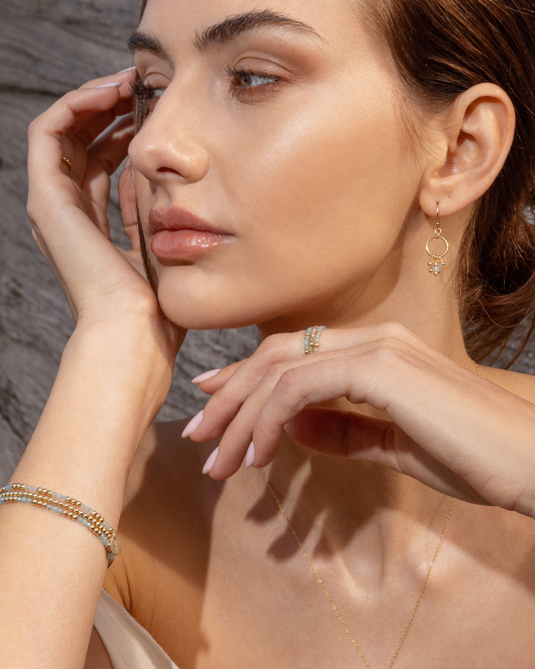 Close-up of a woman wearing gold blue topaz gemstone earrings, apatite gemstone rings, and bracelets with a neutral background