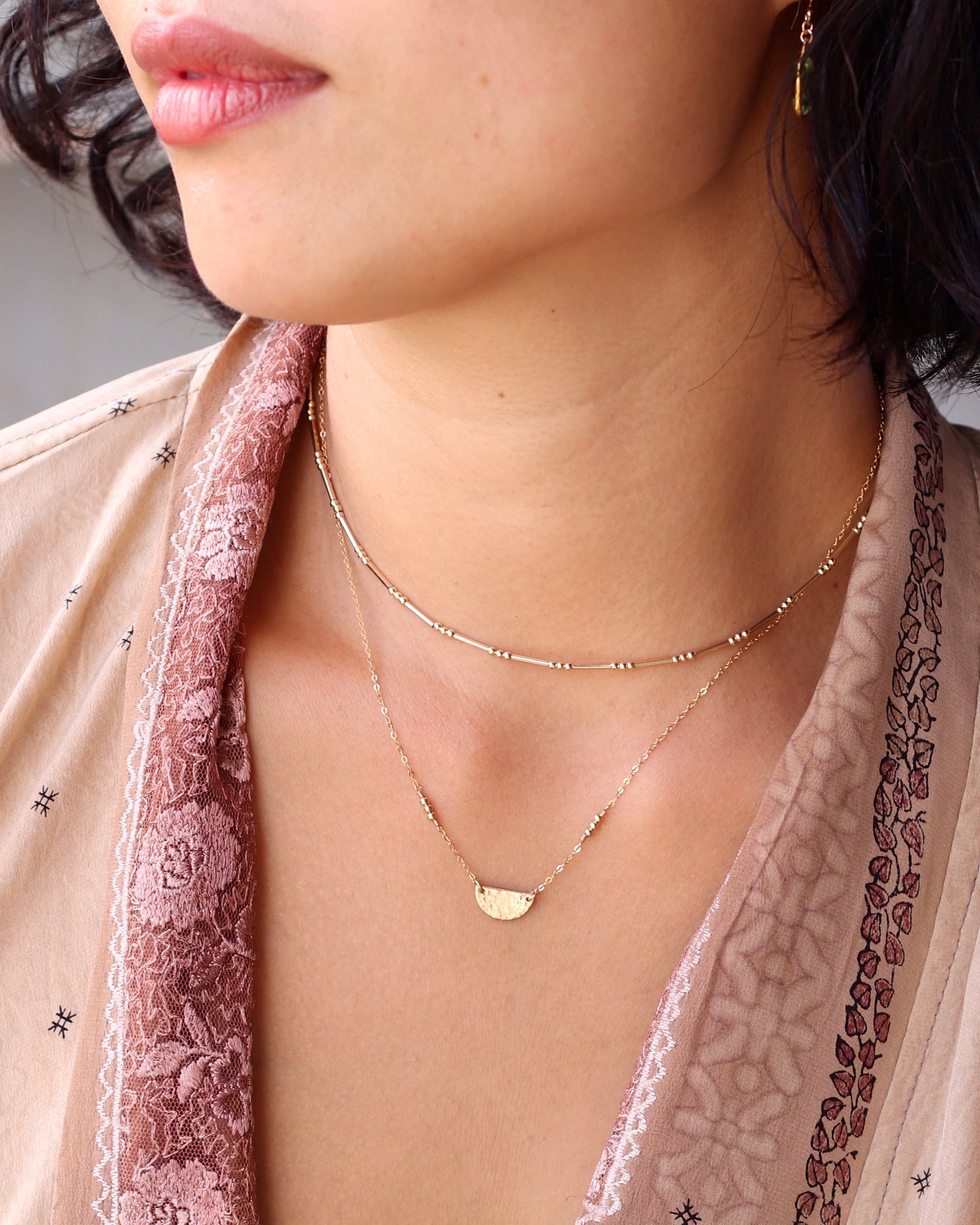 Close-up of a woman wearing a delicate gold necklace with a half circle pendant layered with a gold beaded necklace wearing a pretty floral top