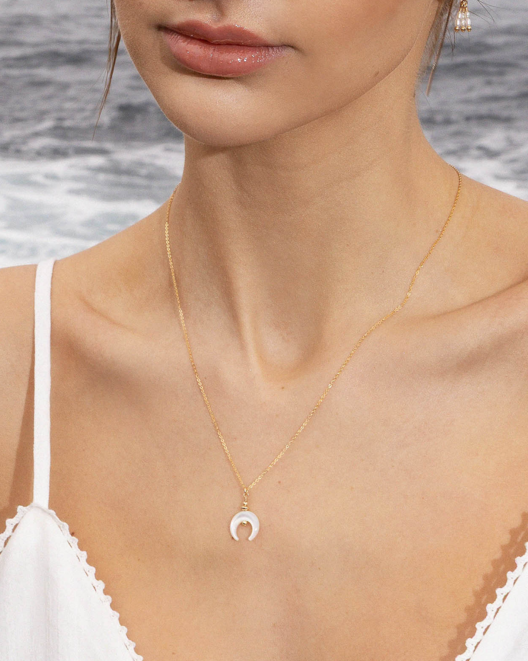 Close-up of a woman's neck wearing a gold necklace with a mother of pearl crescent moon pendant and pearl thread earring by the ocean.