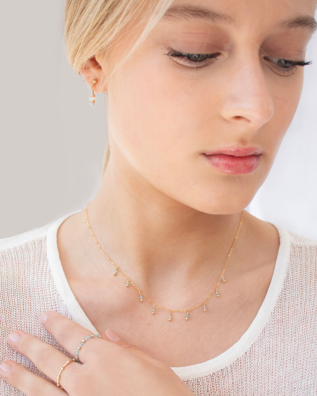Woman wearing a delicate gold apatite gemstone necklace, rings and opal earrings with a plain background