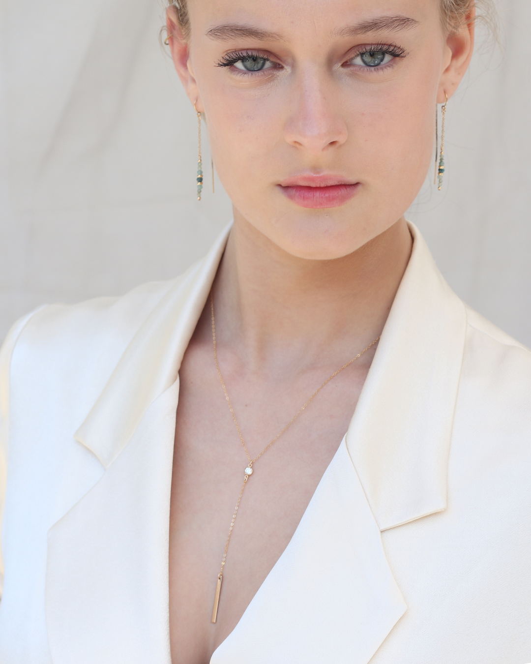 Woman wearing a cream blazer with a gold thread grandidierite gemstone earrings and a gold lariat cubic zirconia necklace against a plain background