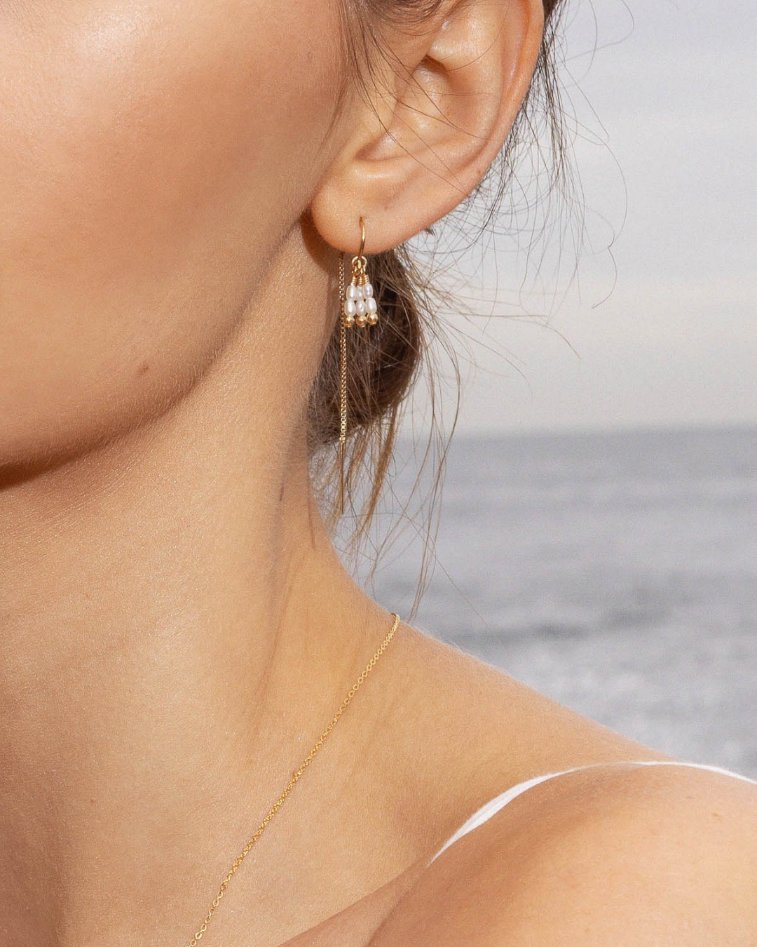 Close-up of a person wearing a gold thread pearl earring against a blurred ocean background