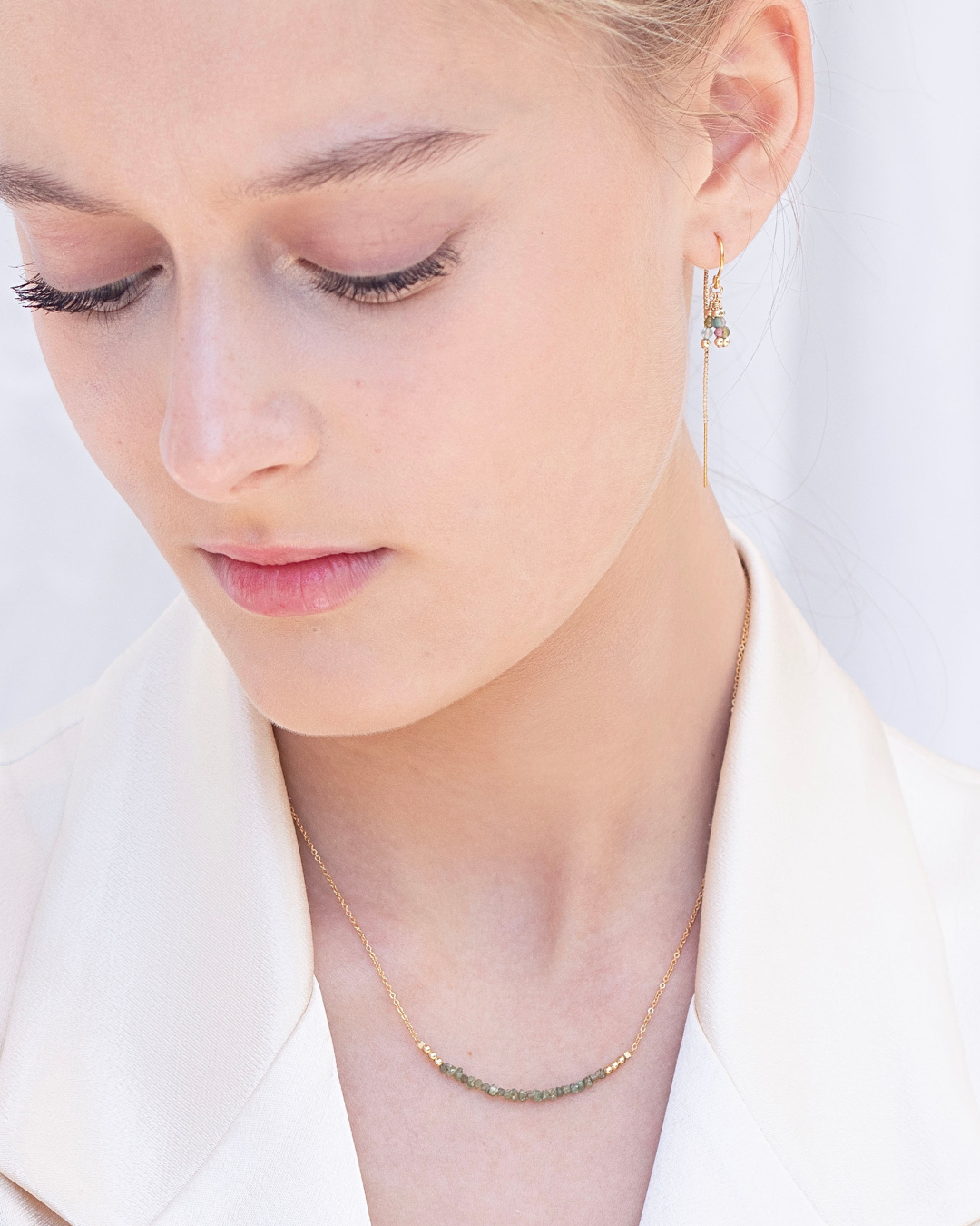 Close-up of a woman wearing a delicate gold necklace with green raw diamond gemstones and a tourmaline thread earring against a light background