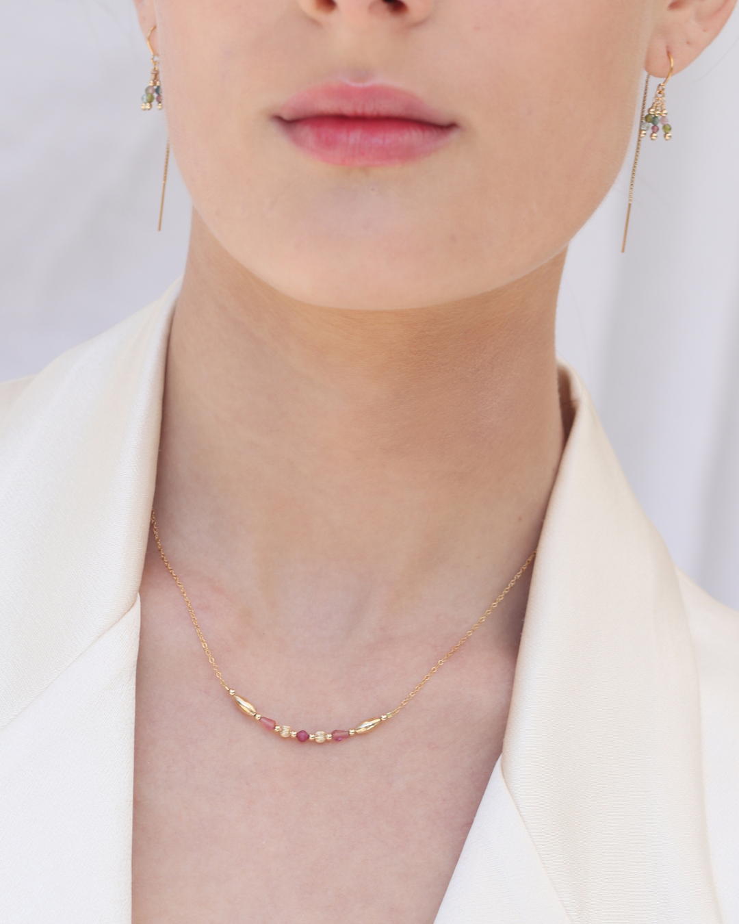 Close-up of a woman wearing gold tourmaline earrings and a gold necklace with pink tourmaline gemstones against a plain background